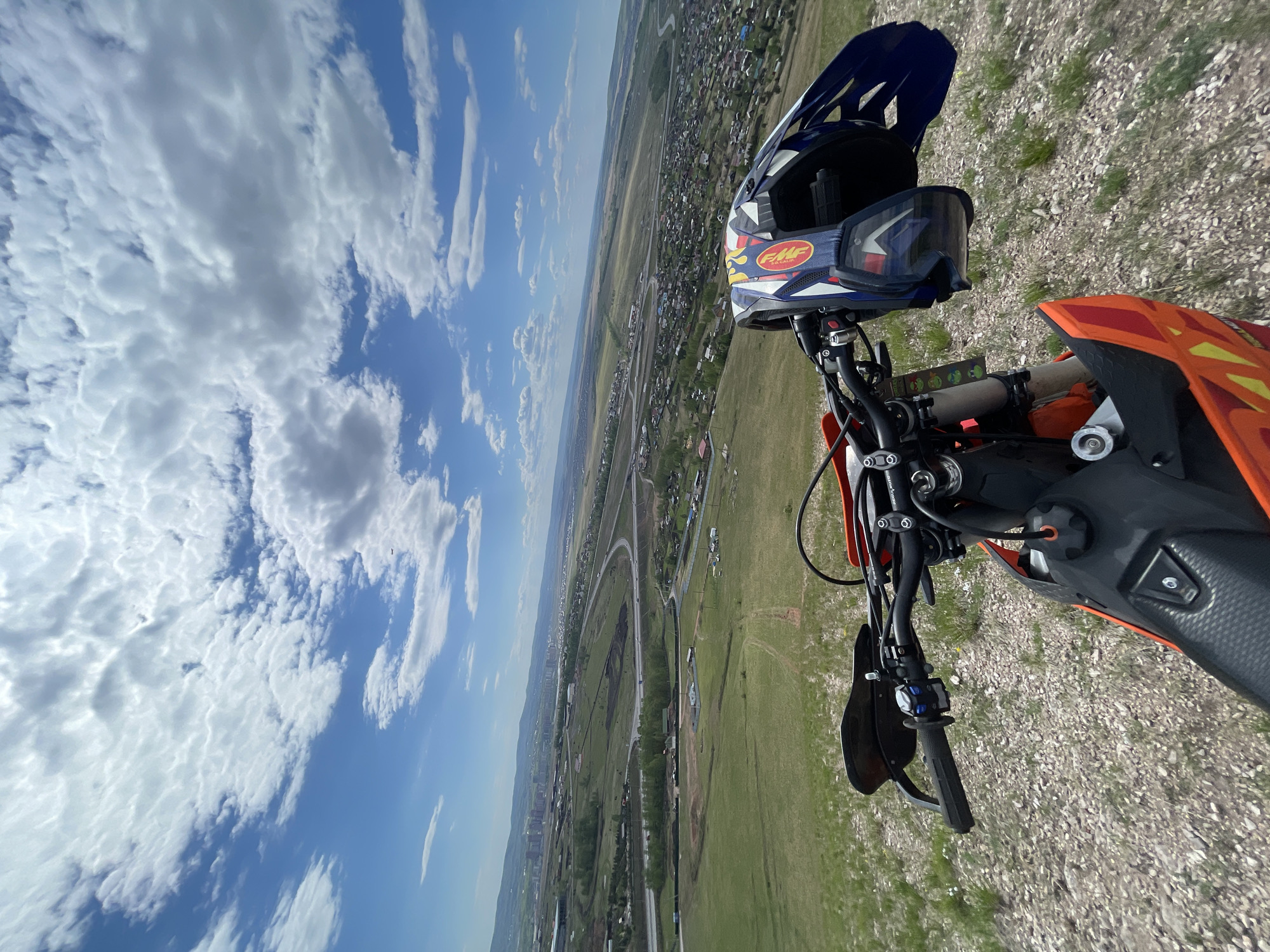 A high-angle, slightly tilted perspective shows the handlebars of a dirt bike and a helmet resting on them. The helmet is dark blue, white, and red with goggles. The bike has orange and black accents. Below the bike, there's a view of Солонцы, Russia, including a network of roads, green fields, and buildings. The sky is mostly blue with fluffy white clouds. The ground beneath the motorcycle appears to be gravel and sparse grass.