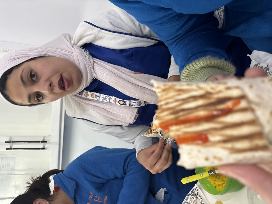 A woman wearing a headscarf and a blue and white zip-up jacket looks towards the camera. She has dark hair and light skin. A child wearing a blue jacket is partially visible. In the foreground, a person holds a square-shaped piece of grilled flatbread drizzled with red sauce. A green bowl with yellow food and a yellow and silver utensil are also in the foreground.