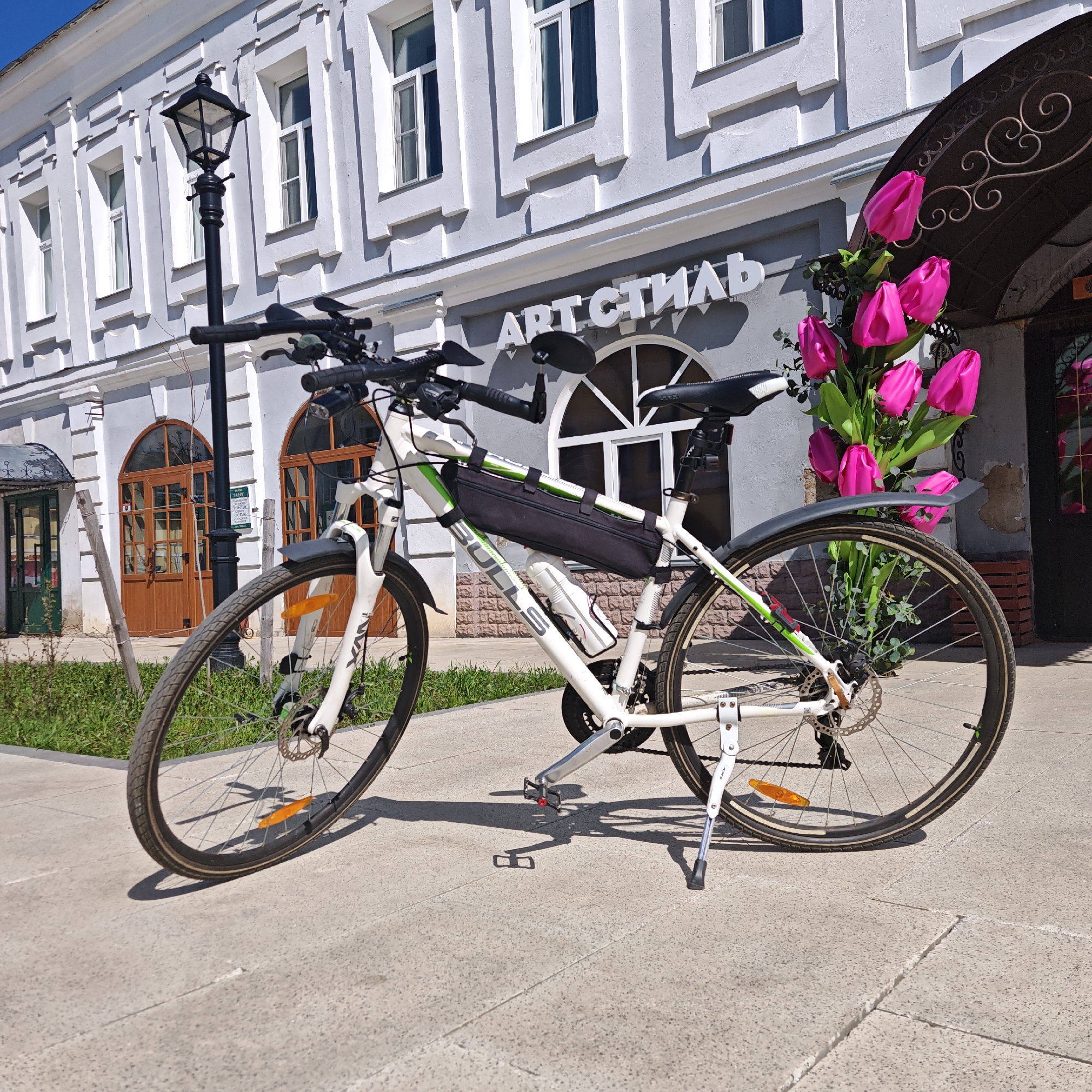 A white Bull's bicycle is parked on a light gray paved sidewalk in front of a light gray two-story building. The bicycle is facing left, with its kickstand down. A black frame bag is attached to the bicycle frame, and a white water bottle is in a cage. Pink tulip-like artificial flowers are behind the bicycle. Above the bicycle is a sign reading "АРТ СТИЛЬ" in white letters. The building has windows with white trim and arched doorways. A black lamppost is to the left of the bicycle. A small patch of green grass is visible to the left. The image was taken in Rostov, Russia.