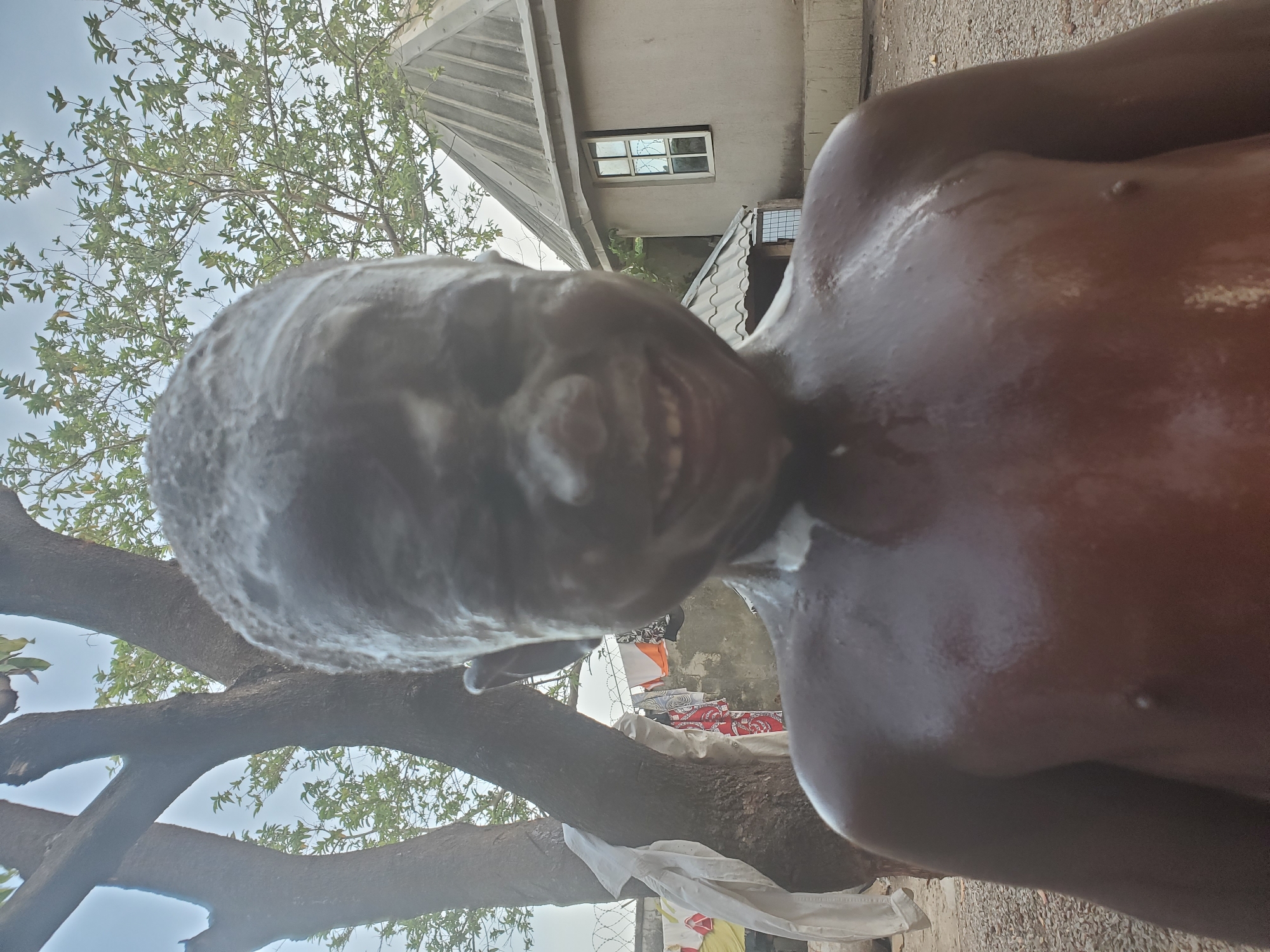 A man with soap or cream covering his face and chest stands outdoors. A tree with leaves is to his left. In the background, a building with a window and a corrugated metal roof can be seen. Clothes hanging on a line and barbed wire are also in the background. The location is likely in Mararaba, Nigeria.