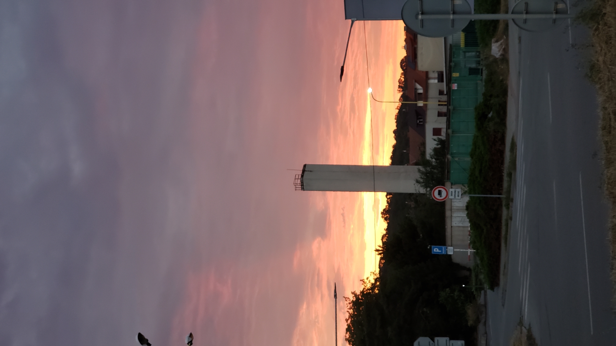 A long, white cylindrical tower stands tall against a dramatic sunset sky painted with hues of pink, orange, and purple. The tower has a square railing structure at its peak. In the foreground, a street corner is visible with a crosswalk. Road signs are present, including a blue parking sign and a circular red and white sign depicting a truck, indicating a truck prohibition. Behind the signs, dense green bushes form a hedge. Several buildings with red-tiled roofs are situated in the middle ground, partially obscured by trees. A streetlamp with a visible light bulb is positioned to the right of the tower. The overall scene suggests an industrial or semi-urban environment at dusk. The specific location in Prešov, Slovakia, is the area around the former ZVL industrial complex, near the intersection of Šebastovská and Jesenná streets.