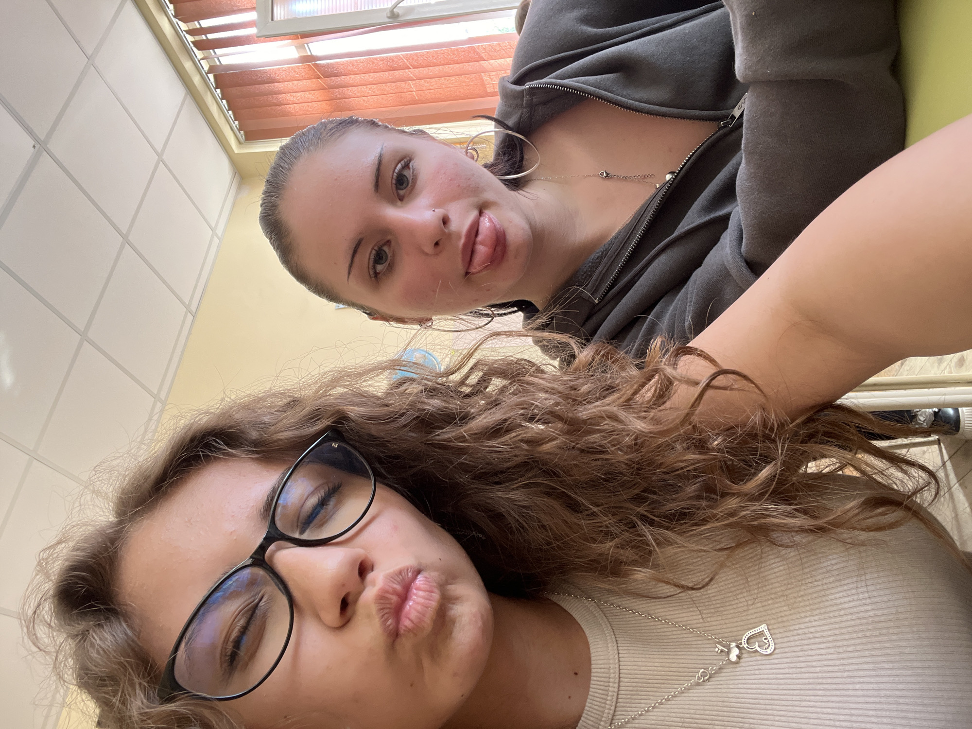 An indoor, low-angle selfie captures two young women lying down and posing playfully. The woman in the upper right, partially obscured, has light skin, dark hair pulled back, large silver hoop earrings, and a nose piercing. Her tongue is slightly out in a playful expression. She wears a dark gray hooded zip-up sweatshirt and a delicate silver chain necklace. The woman in the lower left, closer to the camera, has long, wavy brown hair and black-framed glasses. She makes a pouty or kissing face and wears a light-colored ribbed top with a silver necklace featuring a heart-shaped pendant. The background reveals a white grid-patterned drop ceiling and a window with horizontal brownish-orange blinds, suggesting natural daylight. A small portion of a light green surface is visible in the top right corner. The overall mood is casual and friendly. No text is visible in the image.