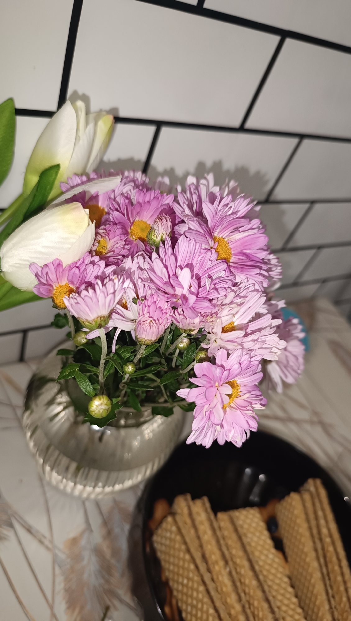 In Chaikovsky, Russia, on a countertop, a bouquet of pink chrysanthemums and white tulips sits in a clear glass vase. To the right of the vase is a black bowl filled with wafer cookies. The background consists of a tiled wall with white tiles and dark grout lines.