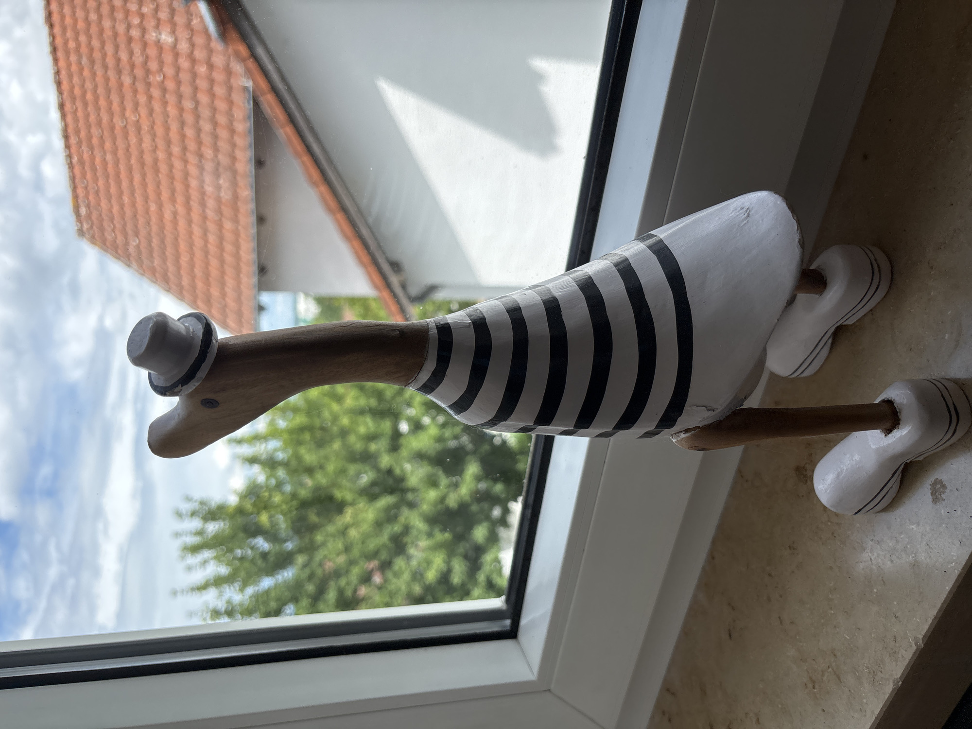 A wooden decorative figure resembling a duck or goose, dressed in a striped shirt and a top hat, stands on an indoor windowsill. The figure's head and neck are made of light brown wood, with a small white top hat featuring a black band affixed to its head. Its body is painted white with horizontal black stripes, while its two slender wooden legs connect to two separate white, shoe-like bases. Minor paint chipping is visible on the figure's body and bases.
The windowsill is made of a light beige or off-white stone material, displaying a subtle texture and minor surface irregularities. Behind the figure, a white window frame with a visible black seal is present. Through the window, an outdoor scene is visible but blurred due to a shallow depth of field. A partly cloudy blue sky dominates the upper portion of the exterior view. Below the sky, out-of-focus green foliage from trees is prominent. To the right, a residential building with an orange-red tiled roof and a white facade is partially visible. A distinct triangular shadow is cast on the white wall of the distant building. The scene is illuminated by natural light, with the indoor elements appearing sharp and the outdoor elements intentionally softened. The location is Unknown, Unknown.