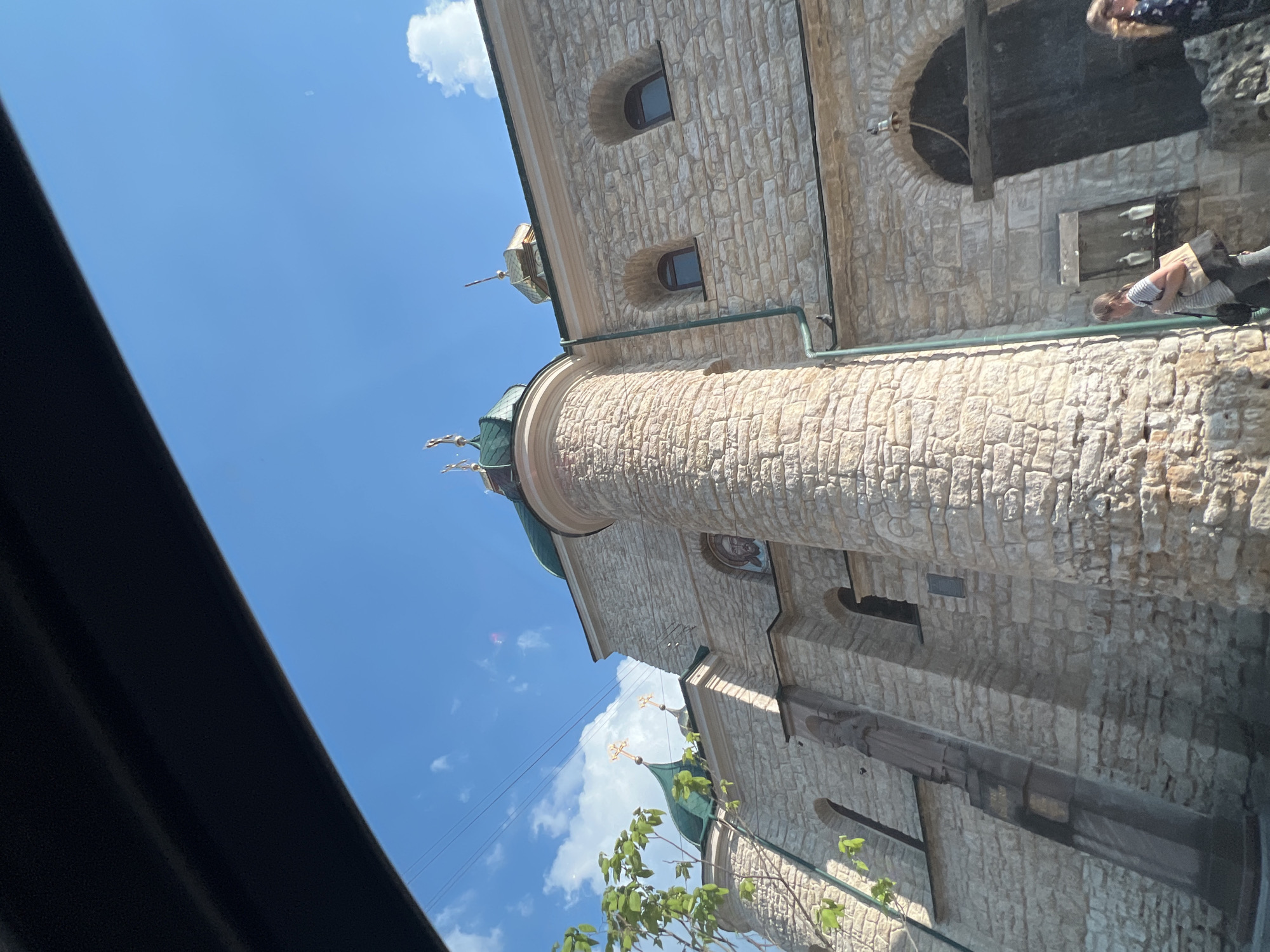 This media file depicts an exterior view of a stone building, likely a church, under a bright blue sky with scattered white clouds. The building's architecture is characterized by its rough-hewn stone construction and arched windows. A prominent round tower with a green-domed roof and a cross adorns the structure. Several other crosses are visible on smaller domes and spires.
In the lower portion of the image, the edge of a dark object, possibly a car roof or window frame, partially obscures the view. A few green leaves from a tree or bush are also visible in the foreground.
The scene appears to be set during the daytime, with bright sunlight casting shadows. Two individuals are partially visible in the image. One person, with blonde hair, is seen from above on the right side of the frame, appearing to be walking or looking down. Another person, wearing a striped shirt and carrying a bag, is leaning down near the building on the right side, possibly interacting with the structure or an object near it.
No visible text is present in the image. The overall impression is a sunny day in Ternopil, Ukraine, with a focus on the architectural details of a religious building and glimpses of people going about their day.