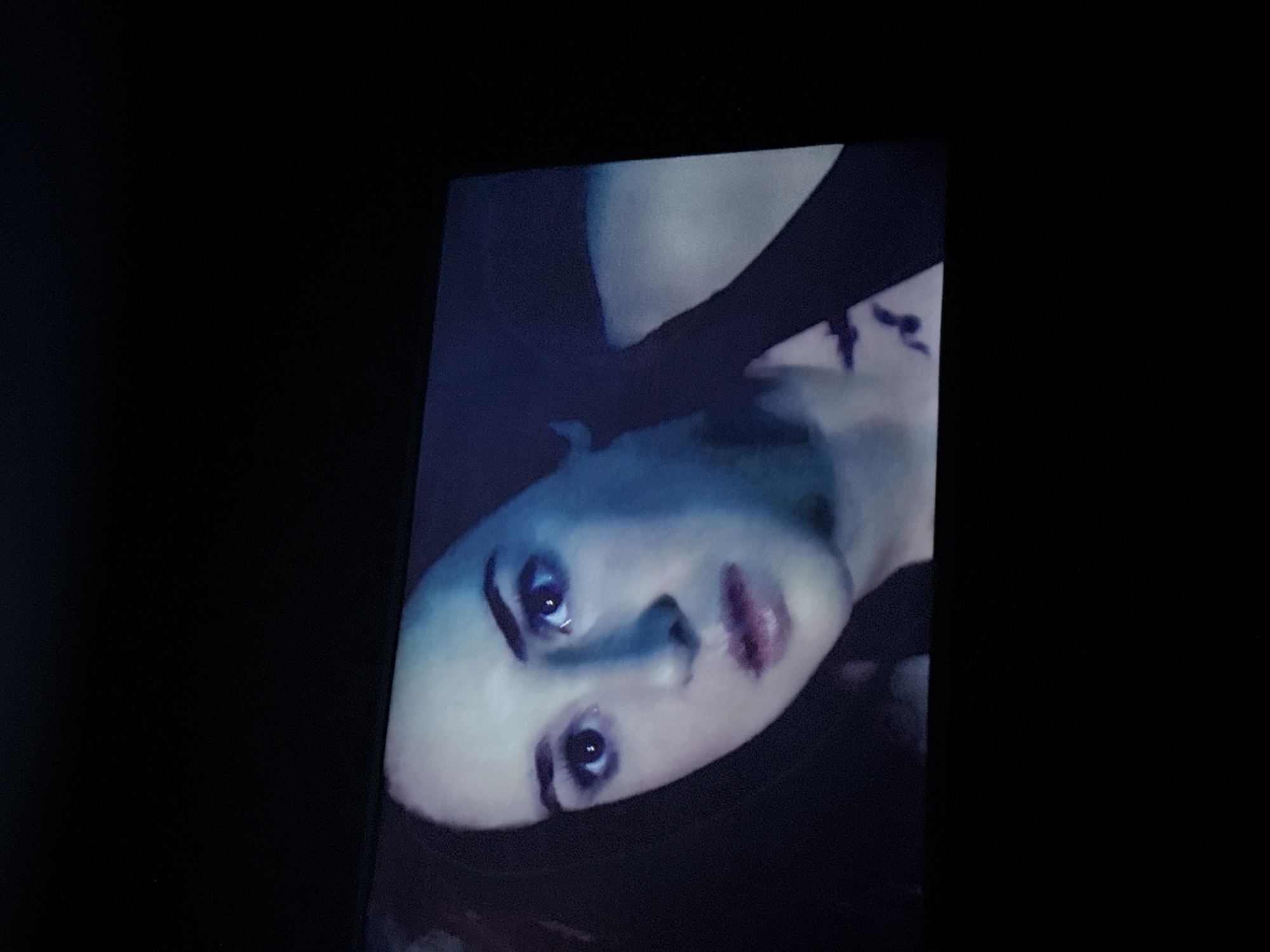 A portrait of a person with light skin, dark hair, and facial tattoos is displayed on a screen in a dark environment. The person has prominent eyebrows, dark eyes, a pointed nose, and dark lips.
The image is off-center and rotated, with the person's face angled towards the viewer. The background is black. The location is not discernible from the image.