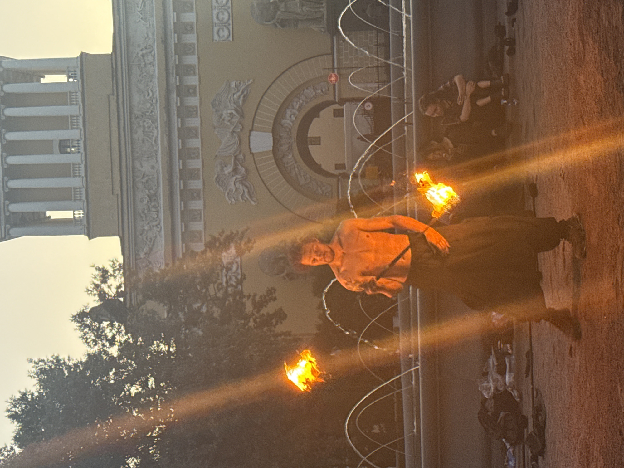 The image depicts a street performance in Saint Petersburg, Russia, likely near the Admiralty Embankment, characterized by the architectural style of the background building. A shirtless male performer, with dark, curly hair and tattoos on his arms, is centrally positioned, engaged in fire dancing. He holds a flaming object in his right hand, and another flame is visible in the air to his left, suggesting he is actively performing. He is wearing dark, flowing pants and athletic shoes.
In the background, a grand classical building with columns, decorative friezes, and arched entrances stands against a pale, dusky sky. In front of the building is a fountain with multiple water jets arcing upwards. The ground appears to be paved or sandy.
To the right of the performer, two individuals are seated on the ground, observing the performance. One person, wearing dark clothing, appears to be smoking. The other, with braided hair, is also seated and looking towards the performer. Scattered around the performance area are various items, possibly personal belongings or props for the performance, including dark fabric items and bags. The scene is illuminated by the natural light of dusk and the bright flames of the performance, with visible light flares streaking across the image.