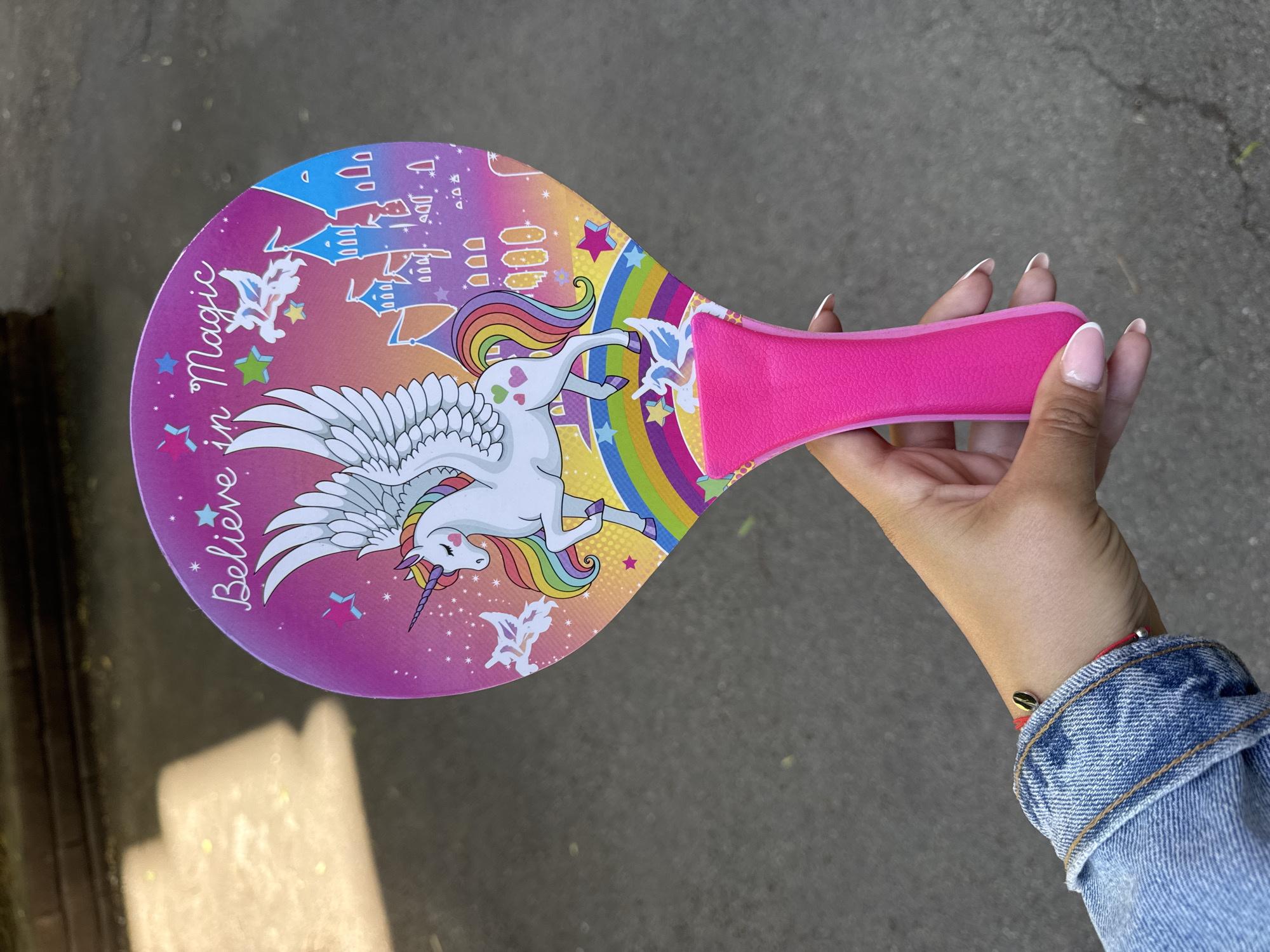 A hand holds a pink unicorn paddle with a "Believe in Magic" message. The paddle has a round shape, a pink handle, and features a unicorn illustration with wings and a rainbow mane, set against a background of a castle, stars, and a rainbow. The hand holding the paddle has a denim jacket on, with a red bracelet visible on the wrist. The background appears to be an outdoor urban setting, specifically a sidewalk. It is not possible to identify Sofia, Bulgaria as the location from this single image.