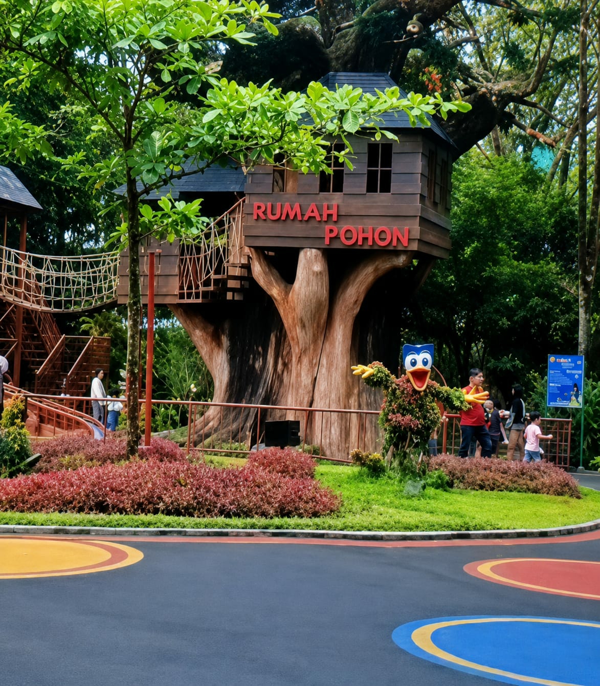 This is a photograph taken in Bandar Baru, Indonesia, featuring a treehouse attraction. The treehouse, labeled "RUMAH POHON," is constructed from wood and built around a large tree trunk. It has a dark roof, wooden walls, and windows. A rope bridge connects to a wooden staircase leading up to the treehouse. In the foreground, there is a paved area with colorful circular markings. Lush green grass and red foliage surround the base of the tree. To the right of the treehouse, a topiary figure shaped like a cartoon character with open arms is situated on a grassy mound. Several people are visible in the background, including children and adults, some interacting near the treehouse and the topiary figure. A blue sign with white text is posted on a pole to the far right. The environment is outdoors with abundant greenery and trees.