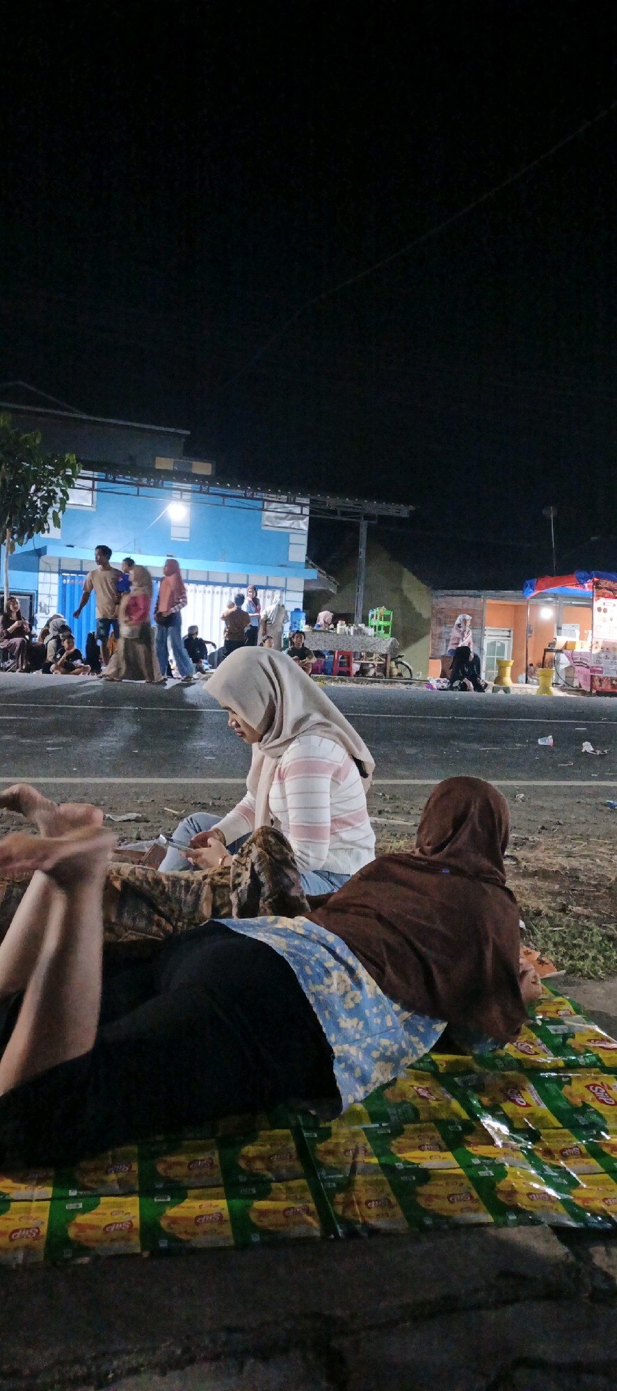 The image depicts an outdoor scene at night, likely in Kasiyan, Indonesia, with several individuals gathered. In the foreground, two people are lying down on a mat made of what appear to be snack wrappers. The person closer to the camera is lying on their back with their legs extended, wearing black pants and a blue and white patterned cloth. A second person is lying on their side, wearing a brown headscarf and a brown outer garment. Partially visible behind them, a person in a light-colored headscarf and a striped shirt is sitting and looking at a phone.
In the mid-ground, a group of people are standing and walking near a building with a blue facade and white trim. Some are wearing headscarves. To the right of the building, there are small stalls or shops with awnings and lighting, suggesting a market or street food area. One stall has colorful displays and yellow stools. The ground appears to be paved or a dirt road. The background is dark, indicating nighttime. The overall atmosphere suggests a casual gathering or a marketplace activity at night.