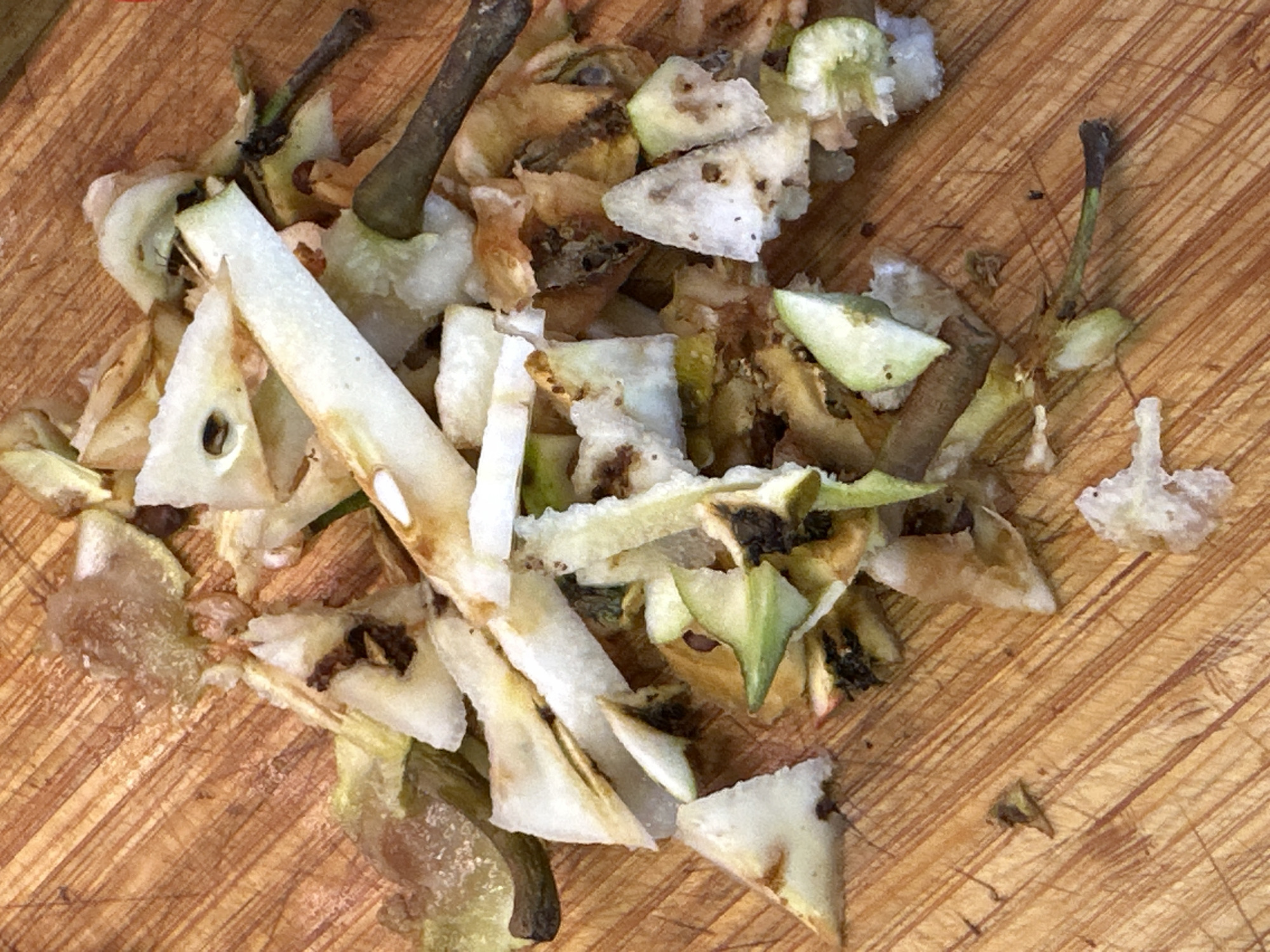 A pile of discarded pear scraps, consisting of skins, cores, and small pieces of flesh, is spread across a wooden cutting board. The pear scraps are predominantly white and light brown, with some green tinges and dark brown seeds visible. The cutting board has a prominent wood grain pattern with visible scoring marks. The image is shot from a top-down perspective. This scene does not provide any information about the city of Nevinnomyssk, Russia.