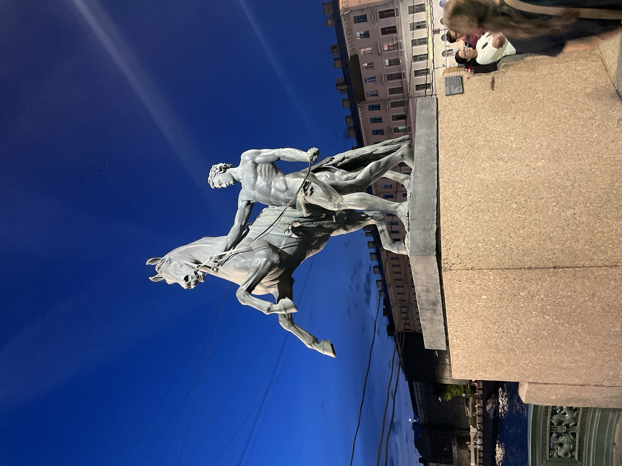 This detailed image captures an evening or night scene on the Anichkov Bridge in Saint Petersburg, Russia, focusing on one of the "Horse Tamers" sculptures. The central subject is a large bronze statue depicting a nude, muscular male figure attempting to control a powerful, rearing horse. The horse is positioned on its hind legs with its front hooves raised high, its head turned slightly to the viewer's left, and its mouth agape. The man, depicted with significant musculature, is shown barefoot and gripping the horse's reins and flank.
The statue is mounted on a tall, textured, light-brown granite pedestal. The sky above is a deep twilight blue, with faint, wispy clouds visible and a distinct white contrail stretching diagonally from the upper left towards the upper right. Several dark, thin wires or cables are strung across the sky, particularly prominent on the left side of the frame.
In the lower left, a section of a green, ornate cast-iron bridge railing is visible, featuring intricate decorative elements that resemble mythical creatures. Below this railing and to the left of the pedestal, the dark waters of the Fontanka River are visible, reflecting ambient lights. Further in the background on the left, dark buildings and structures, some partially obscured by scaffolding, line the riverbank. On the right side of the frame, behind and to the right of the pedestal, stands a light-colored, multi-story building with numerous windows, some of which are illuminated, indicating internal activity.
Three individuals are visible in the lower right background, behind the statue's pedestal. One woman, wearing a light-colored top and glasses, stands with her arms crossed, looking towards the left side of the frame. Partially obscured behind her is a man in a darker shirt. Closest to the viewer on the far right, only the back of another person's head and shoulder are visible, indicating they are moving or positioned with their back to the camera. The scene is illuminated by artificial light sources highlighting the statue against the dark sky and providing ambient light to the surrounding environment.