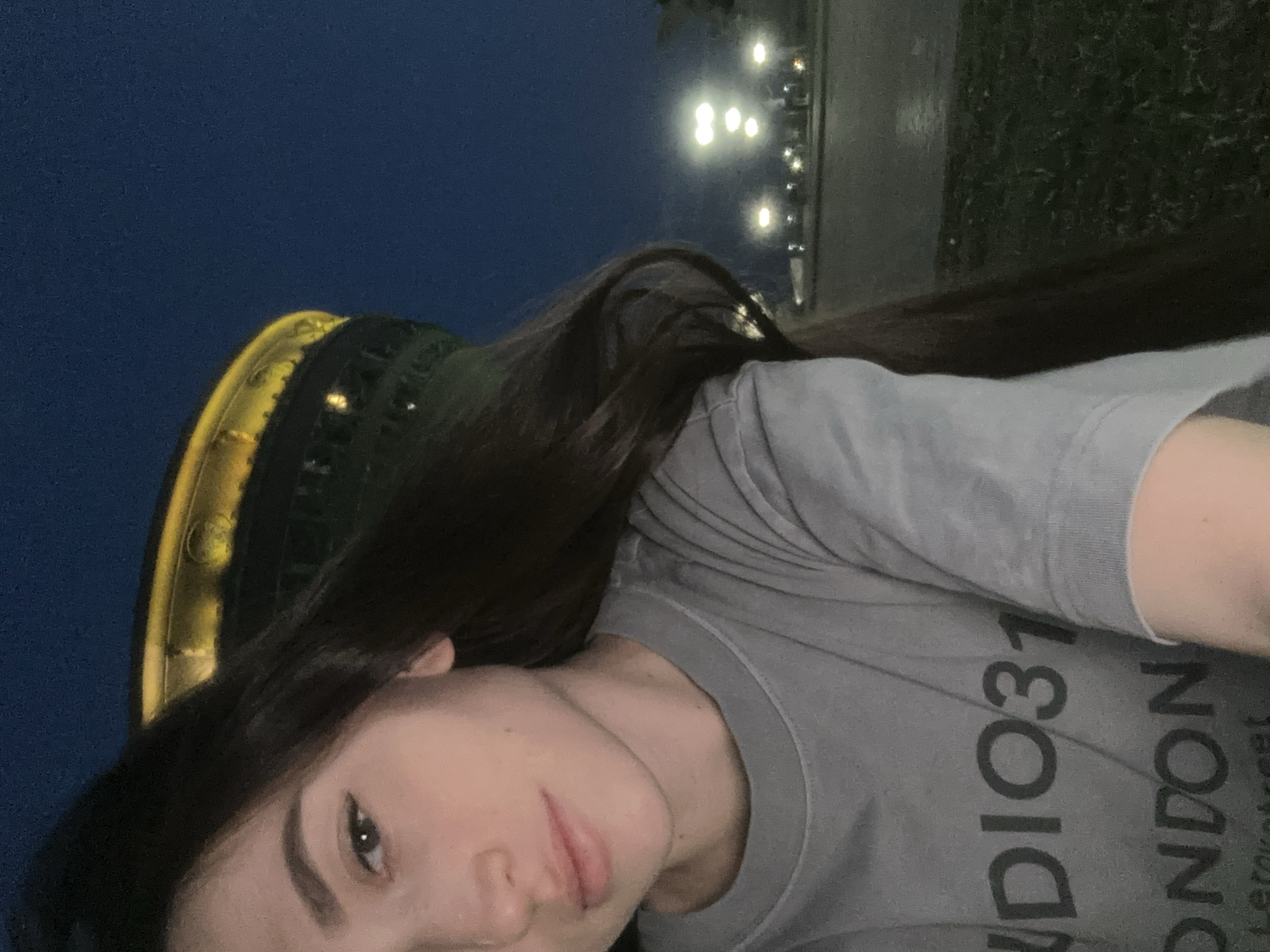 A selfie shows a woman with dark hair wearing a gray t-shirt with "STUDIO31 LONDON" printed on it. In the background, on the left, is the Qolsharif Mosque in Kazan, Russia. Street lights are visible in the background. The sky is dark. The woman is facing the camera.