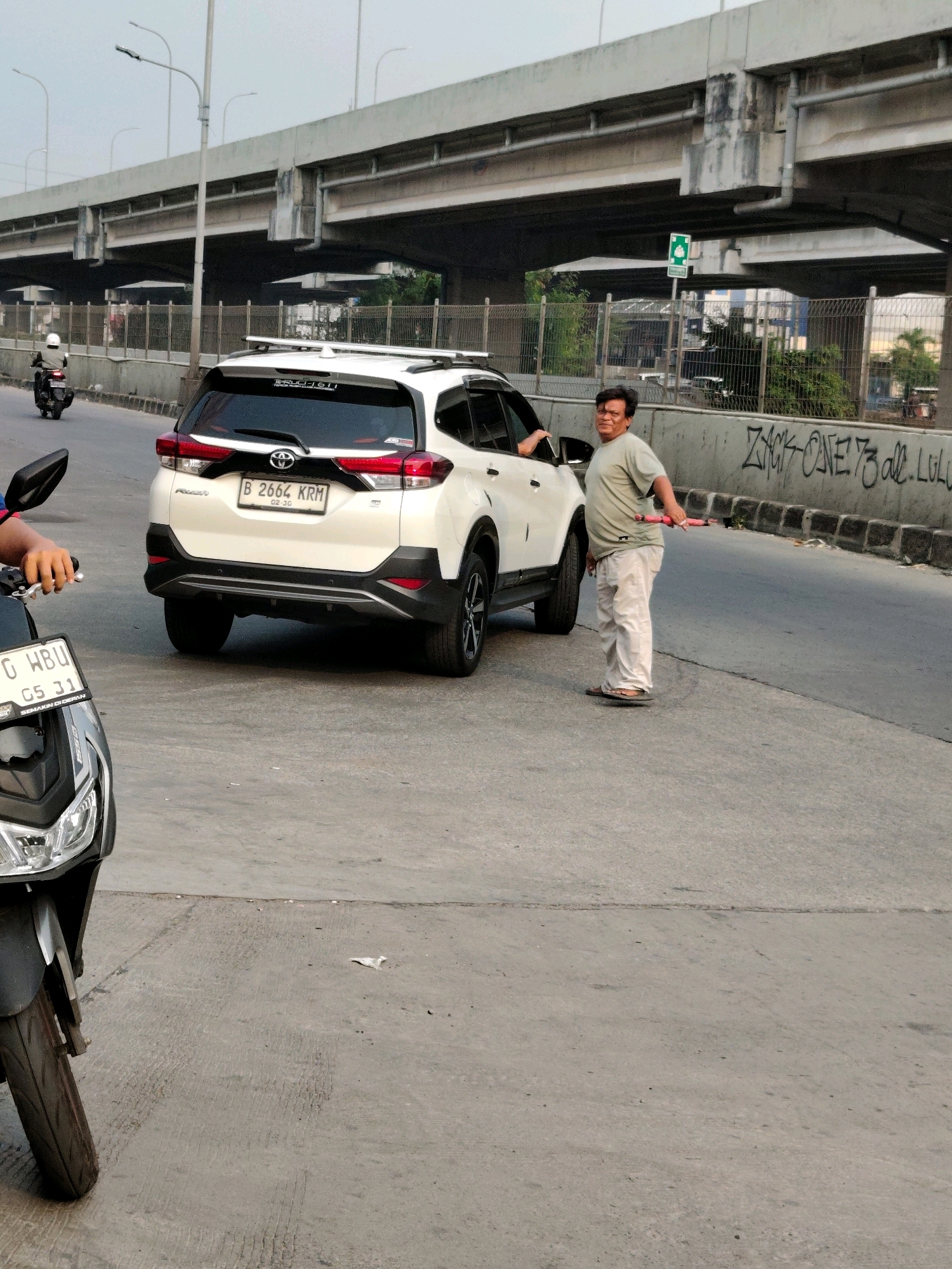 A middle-aged man wearing a light-colored t-shirt and light-colored pants stands on the right side of a white SUV, holding a red and white striped pole. The man is looking towards the camera. The SUV has its driver-side door open and the license plate reads "B 2664 KRM". A portion of a dark-colored scooter is visible in the foreground on the left side of the image, with its front wheel and headlight showing. The scooter's license plate is partially visible and reads "G WBU 05 31". The scene appears to be on a road under an elevated highway structure. The highway pillars and supporting beams are visible above. The environment suggests an urban setting in Bekasi, Indonesia, with a concrete road and a concrete barrier wall on the right side of the road, which has graffiti on it. There is also some greenery visible in the background, behind the barrier wall. The sky is overcast.