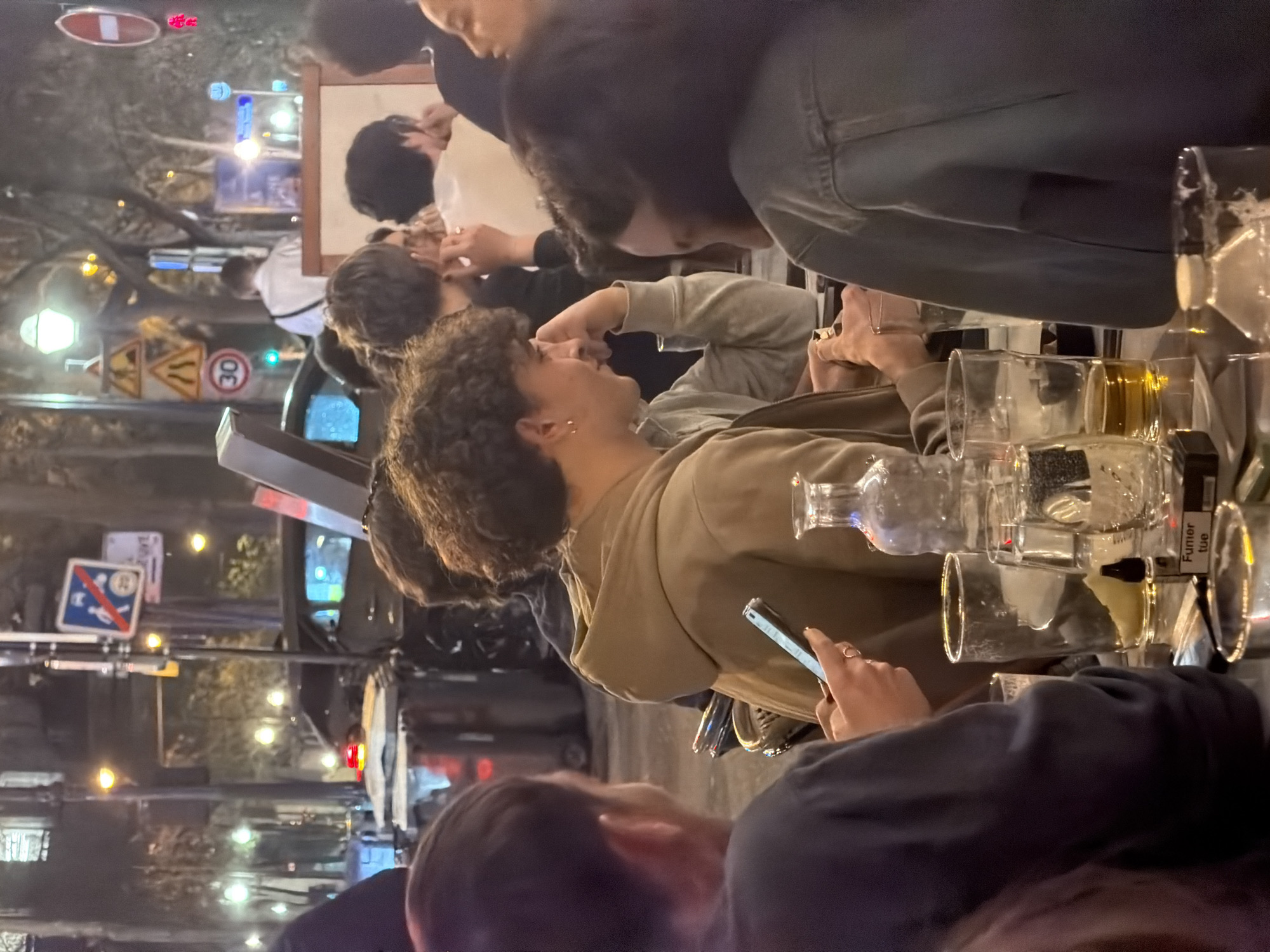 The image captures an outdoor dining scene in Paris, France, likely in the evening or nighttime based on the lighting. Several people are present at the table, engaged in conversation and using their phones. Empty and partially filled glassware, including beer glasses and carafes, are visible on the table. Street signs, including a pedestrian crossing sign and a speed limit sign, are visible in the background. The overall setting appears to be a lively urban environment.