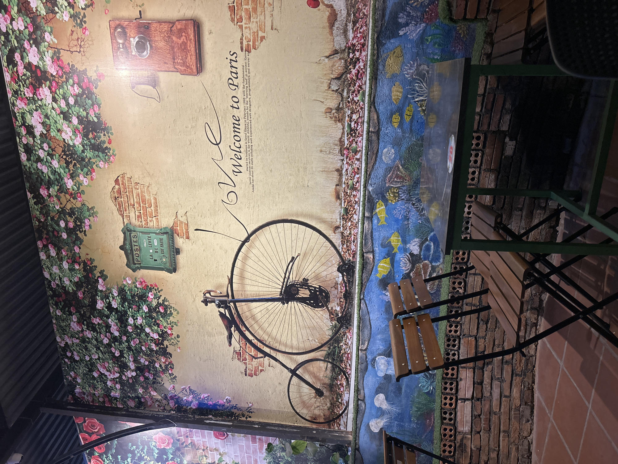 This image depicts an interior wall design in a cafe or restaurant, featuring a composite scene evoking a Parisian theme. The wall is decorated with murals including: floral arrangements in shades of pink and white, a vintage-style bicycle with one large wheel and one small wheel, a painting of an antique-style post box, and a faux brick facade. Additionally, it has a slogan "Welcome to Paris". A table with a marine-themed design is located in front of the wall, along with chairs in front of it. The location is likely in the city of Nha Trang, Vietnam.