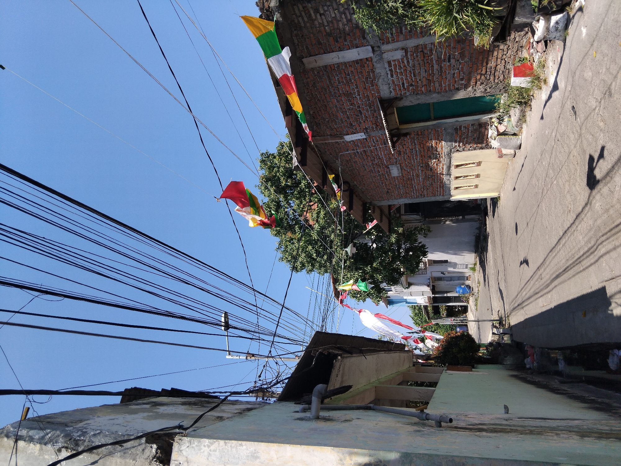 The image depicts a narrow, sunlit street in what appears to be a residential area of Berbek, Indonesia. The street is paved and shows signs of wear. Overhead, a dense network of power lines crisscrosses the sky, casting shadows on the ground. Several colorful banners, likely for a celebration, are strung across the wires and between buildings.
On the left side of the street is a light green building with a weathered facade. To the right, a brick building with a rusted corrugated metal roof is visible, featuring a green wooden door. The architecture suggests a functional, possibly older, urban or semi-urban environment. Scattered debris and some vegetation are present along the edges of the street and in front of the buildings. The sky is clear blue, indicating a sunny day. No people are visible in the scene. The overall impression is of a bustling, lived-in neighborhood.
