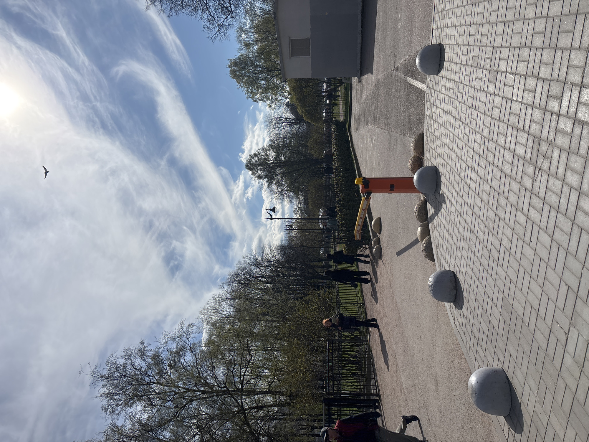 The image depicts a street scene in Saint Petersburg, Russia on a sunny day with wispy clouds in a blue sky. A bird is flying in the sky. Along the side of a paved area, there is a line of concrete bollards of varying sizes. A section of the paved area is constructed of rectangular brick tiles. Adjacent to the paved area is a road or path. Several people are walking along the path. A metal fence separates the path from a grassy area with trees. A building with a window is visible in the background. There's also a yellow and orange parking barrier visible.
