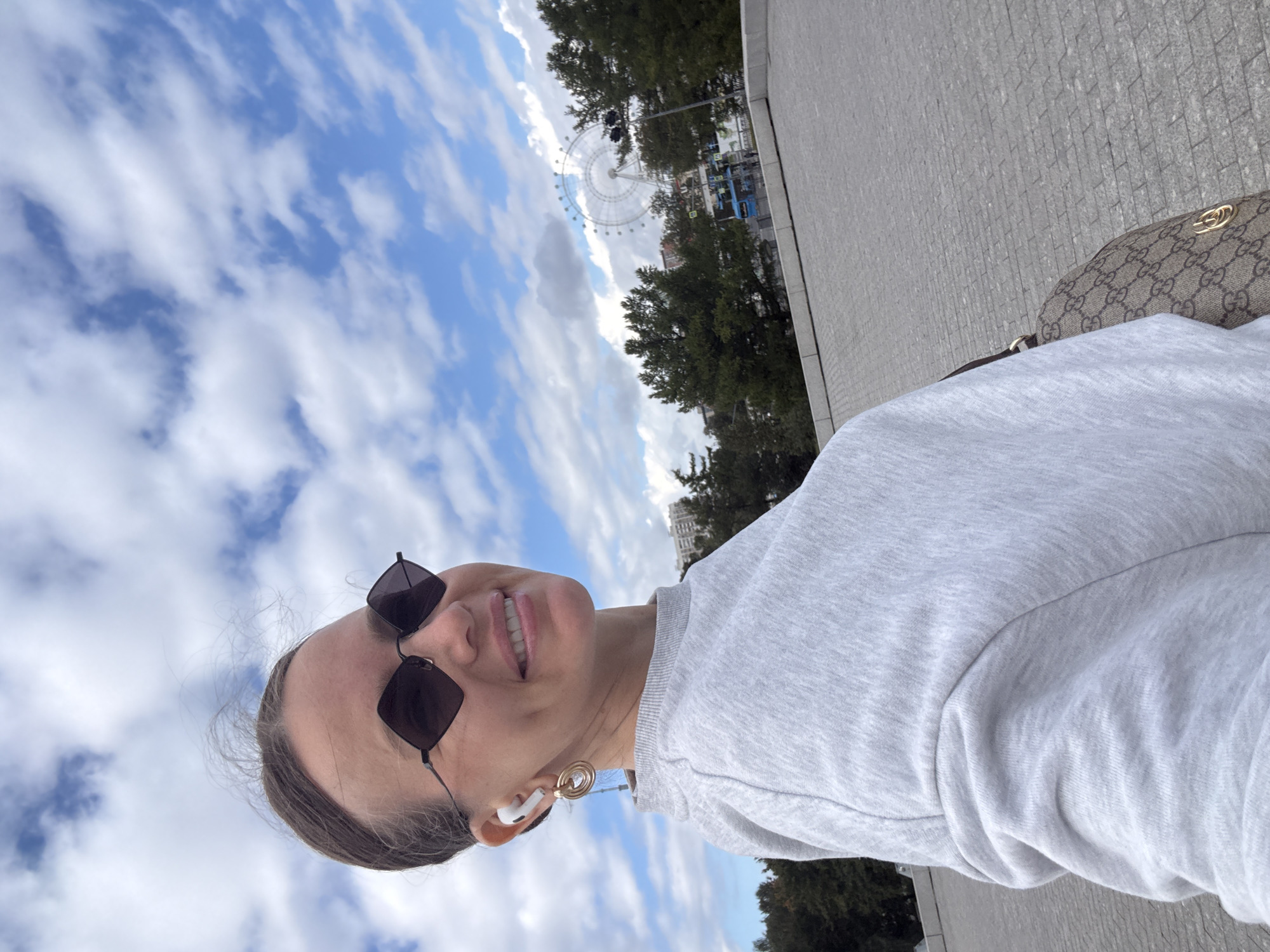 A woman is taking a selfie outdoors in front of a Ferris wheel. She is wearing sunglasses and a gray sweatshirt. She is also wearing white wireless earbuds and gold hoop earrings. She has a brown handbag with a monogram print. The Ferris wheel is in the background, behind some trees. The sky is blue with some white clouds. The ground is paved with gray stones. The location is Moscow, Russia.