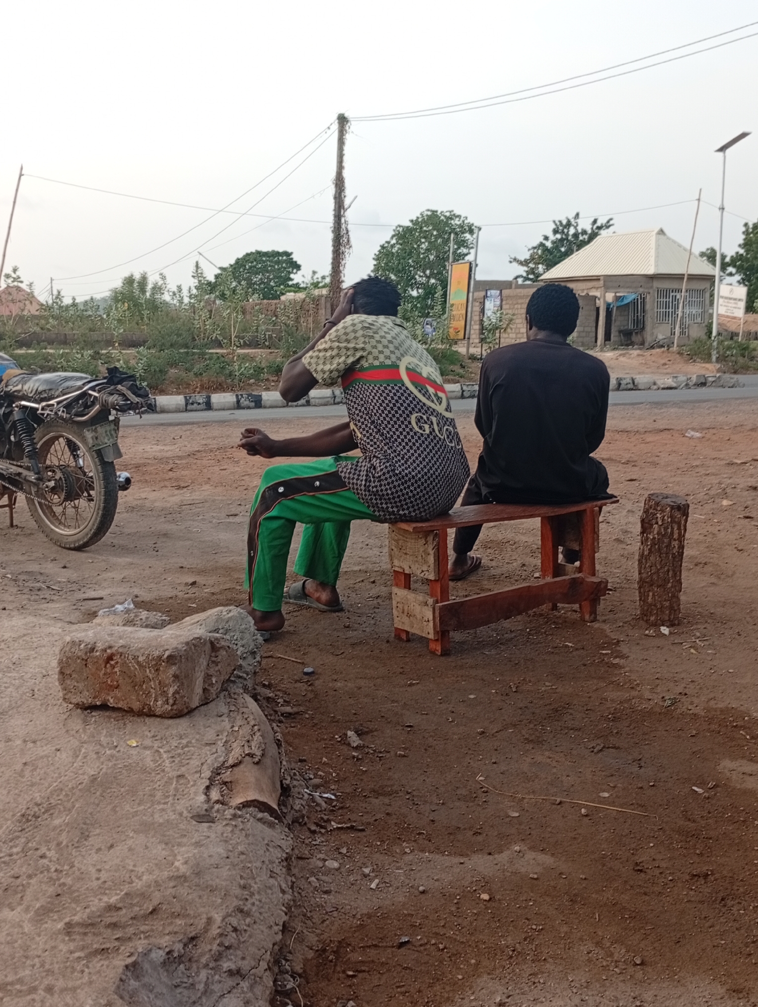 Two men are seated on a small, weathered wooden bench near a road in what appears to be Adankolo, Nigeria. The man on the left wears a patterned shirt, bright green pants with buttons, and sandals. He is gesturing with his hands. The man on the right wears a black shirt and pants, also with sandals. Both men are facing away from the camera. A motorcycle is parked nearby, and a small building with a white roof is visible in the background. The ground is dirt, and there's a stone or brick on the left side of the frame. A cut log rests near the bench, adjacent to the men. Power lines are strung overhead.