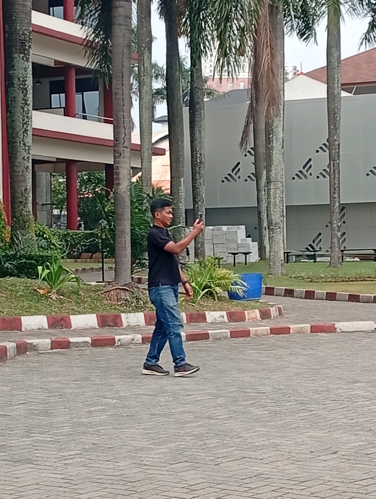 The image shows a man walking on a paved area with a red and white border. He is wearing a black t-shirt and blue jeans, and is holding a phone in his right hand. The background features palm trees, a building with red and white trim, and a modern building with a patterned facade. The scene appears to be an outdoor campus or public space in Kelapa Dua, Indonesia.