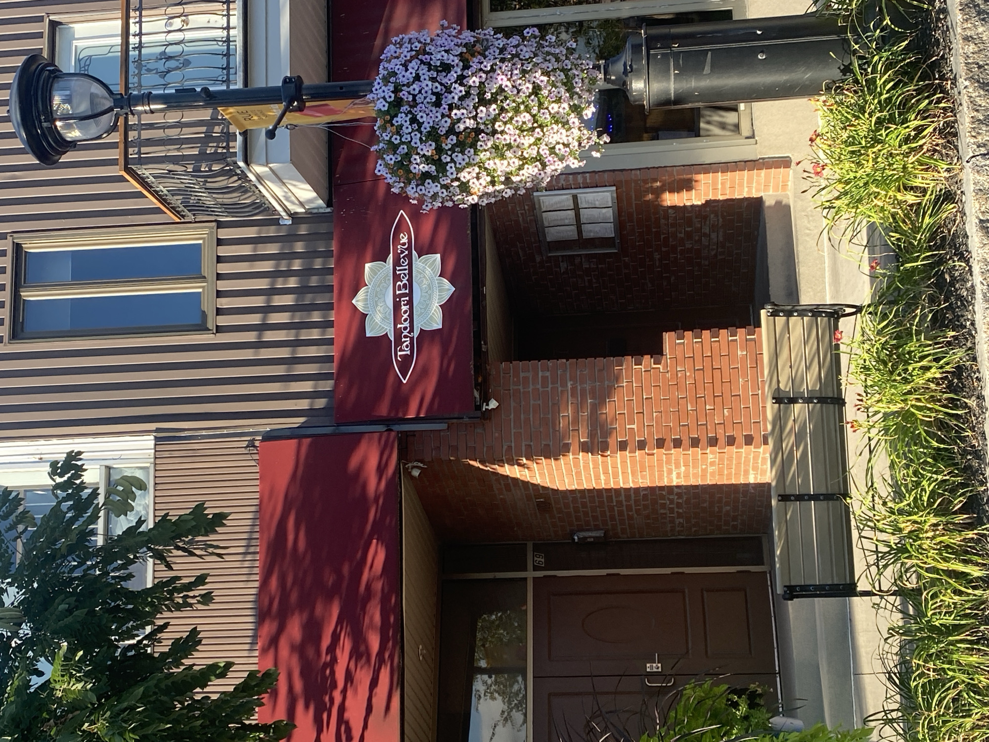 The image depicts the exterior of a restaurant named "Tandoori Bellevue" located in Sainte-Anne-de-Bellevue, Canada. The restaurant has a red awning over the entrance and is constructed with red brick. Above the awning, there is a vertical brown striped facade with windows. A black lamppost with a hanging basket full of white and pink flowers stands to the right of the entrance. The lamppost has a sign attached to it with yellow and red colors. In front of the entrance, there is a brown bench. Lush green plants and some small red flowers are visible in the foreground, with dark mulch covering the ground. The scene is bathed in sunlight, casting shadows from the foliage onto the awning. The architectural style suggests a commercial building in a town or city setting.