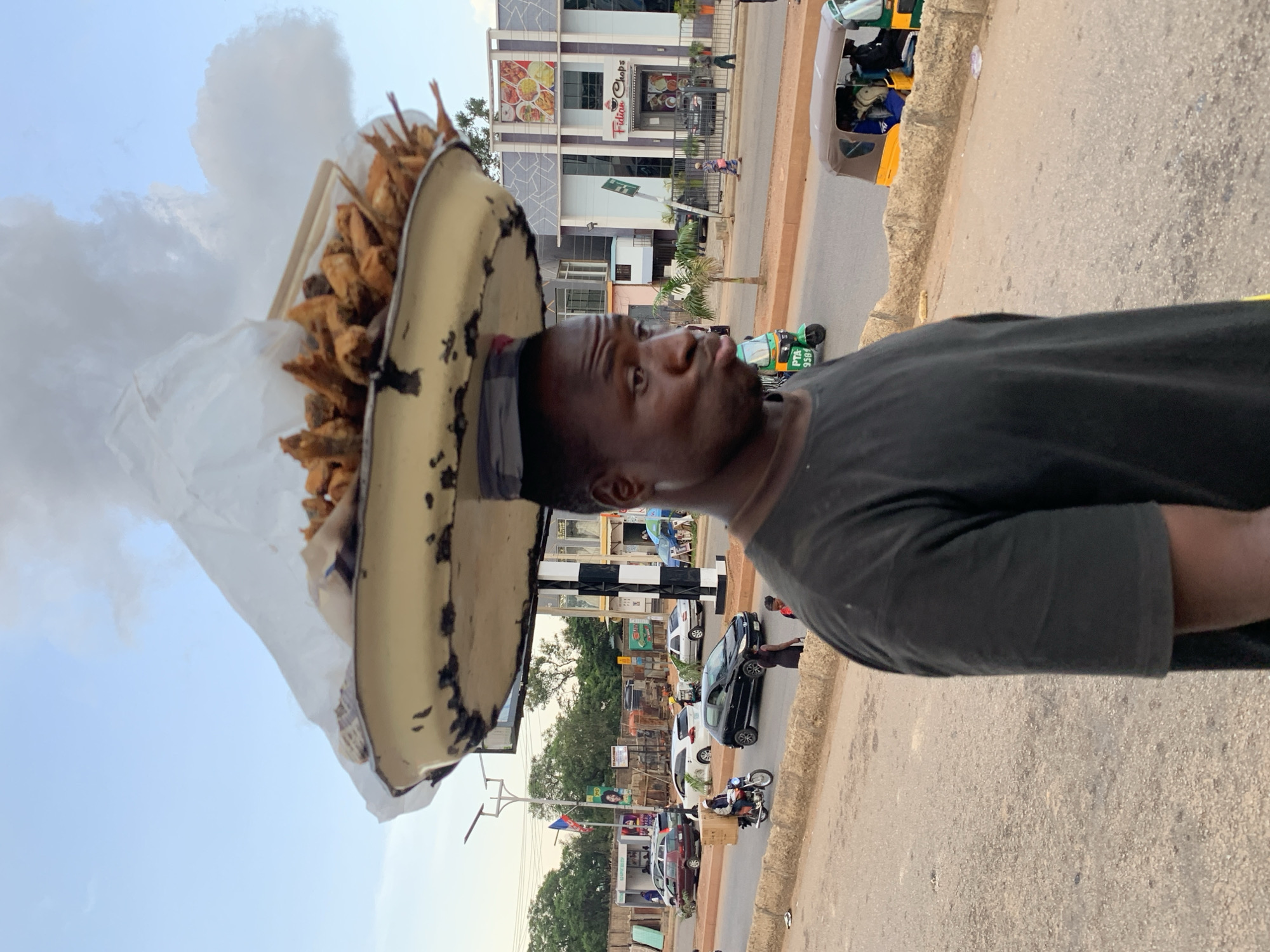A Black man in a black t-shirt carries a wide, shallow metal tray filled with fried fish on his head. The tray is balanced on a flat circular object, possibly a cushion, on his head. The fish appears to be small and whole, and some are sticking out of the tray. A large piece of white plastic sheeting is draped over the top of the fish. The man is in profile, facing left, with his mouth pursed and his eyes looking towards the right.
The scene is a street in Jos, Nigeria. In the background, there is a modern building with "Fidiam Chops" signage and an image of food on its facade. A street with traffic is visible. Cars, a motorcycle with a rider wearing a white helmet and jacket, and a tricycle (keke napep) are present. The sky is partly cloudy. The road surface is asphalt and appears worn. There is a low concrete barrier along the side of the road. The overall lighting suggests it is late afternoon or early evening.
