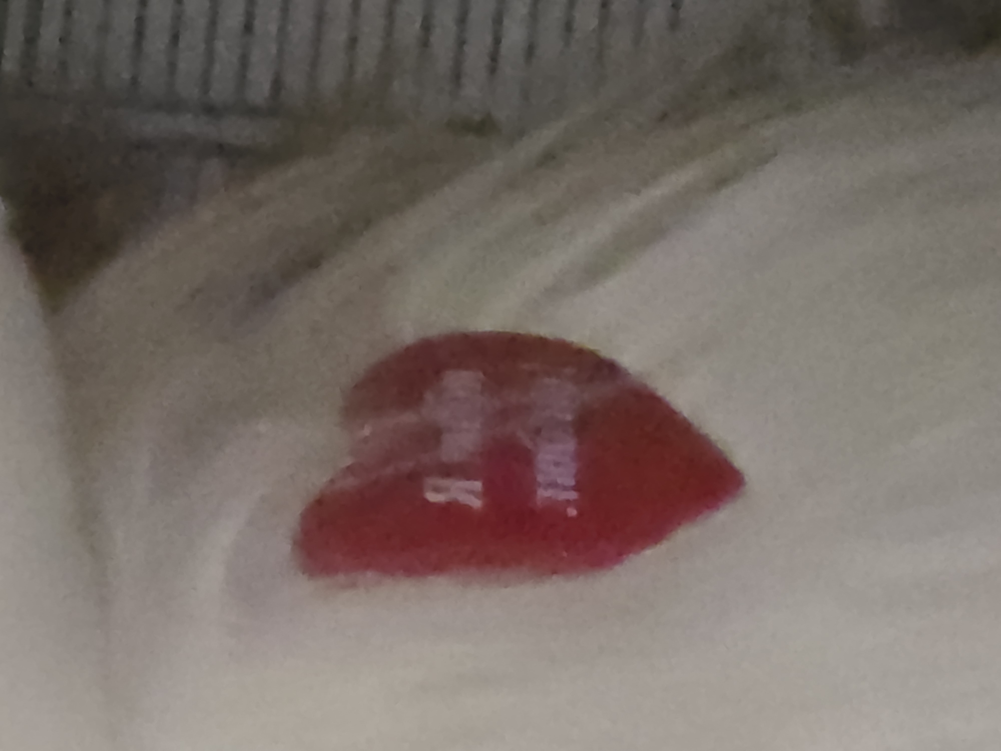 A close-up, blurry photograph shows a red heart-shaped sticker with text on a pale, textured surface. The text on the sticker appears to be in a foreign language, possibly Cyrillic. The sticker is positioned on the left side of the frame. The background is out of focus and appears to be some kind of fabric or textured material with horizontal lines on the right side. The lighting is diffused. The image is not clear enough to determine the exact location, subject, or context.
