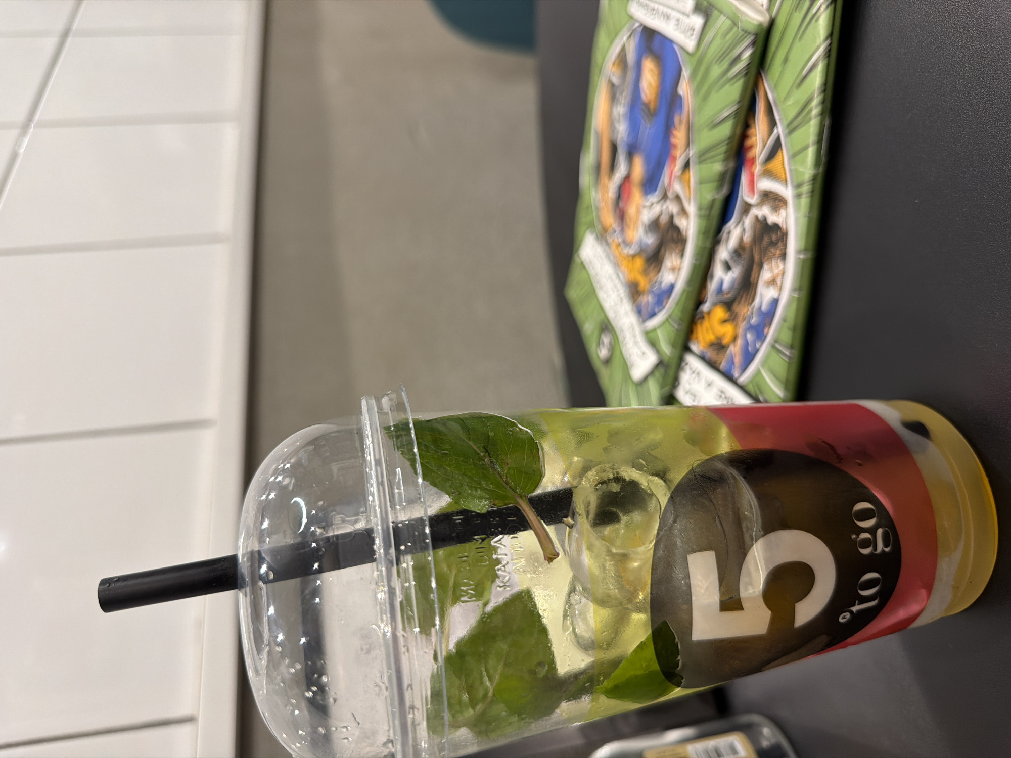 An iced beverage in a clear plastic cup with a domed lid and a black straw is prominently displayed in the foreground. The drink appears to be layered with red and yellow liquids at the bottom, topped with ice cubes, mint leaves, and a light yellow liquid. A black circular sticker with a white number "5" and the text "'to go" is affixed to the side of the cup. In the background, out of focus, are two green pamphlets with colorful illustrations, stacked on a dark surface. The overall setting suggests an indoor environment, possibly a cafe or food establishment in Chișinău, Moldova, given the context of the query. No people are visible in the frame.