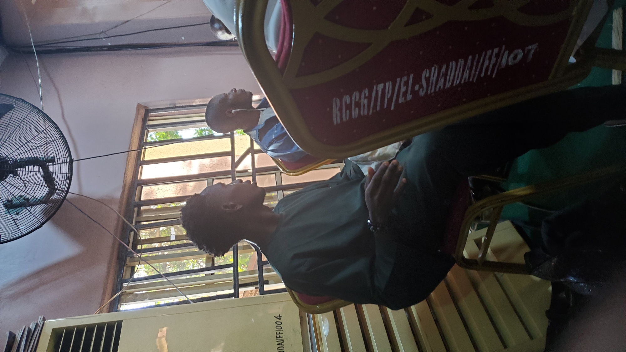 In Jalingo, Nigeria, two young men are seated in chairs, facing towards the right of the frame, in what appears to be an indoor gathering or meeting. The man in the foreground, wearing a dark green traditional garment, is seated on a gold-framed, maroon chair with intricate gold patterns. He has short, textured hair and is looking to his right with his hands clasped loosely in his lap. A black beaded bracelet is visible on his left wrist. To his right, a second young man, wearing a blue and white striped shirt and earbuds, is also seated on a similar chair. He is looking forward and slightly to his right. Behind them, a large industrial fan is mounted on the wall, and a window with vertical bars allows natural light to enter the room, illuminating the dusty air. Adjacent to the window is a tall, beige air conditioning unit. The floor is covered with a dark green material. On the backrest of the chair in the foreground, white lettering is visible, reading "RCCG/ITP/EL-SHADAI/FF/107".