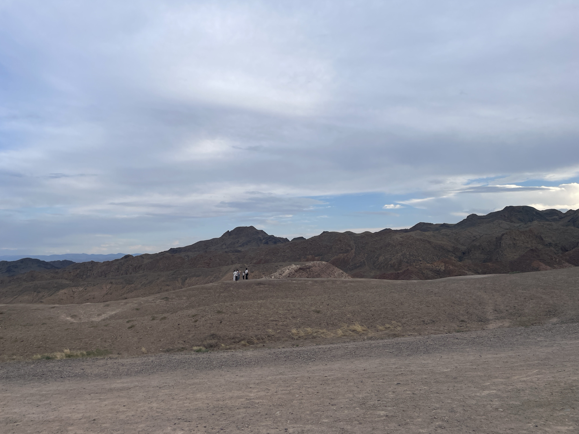 The image depicts a vast, arid landscape with rolling hills and mountains under a cloudy sky. In the middle ground, a small group of four people, appearing to be tourists, are standing on a hilltop. The foreground consists of a dirt path, typical of a rural or natural setting. The location is identified as Tamerlik, Kazakhstan, suggesting a natural park or geological site. The overall impression is one of remote, rugged beauty.