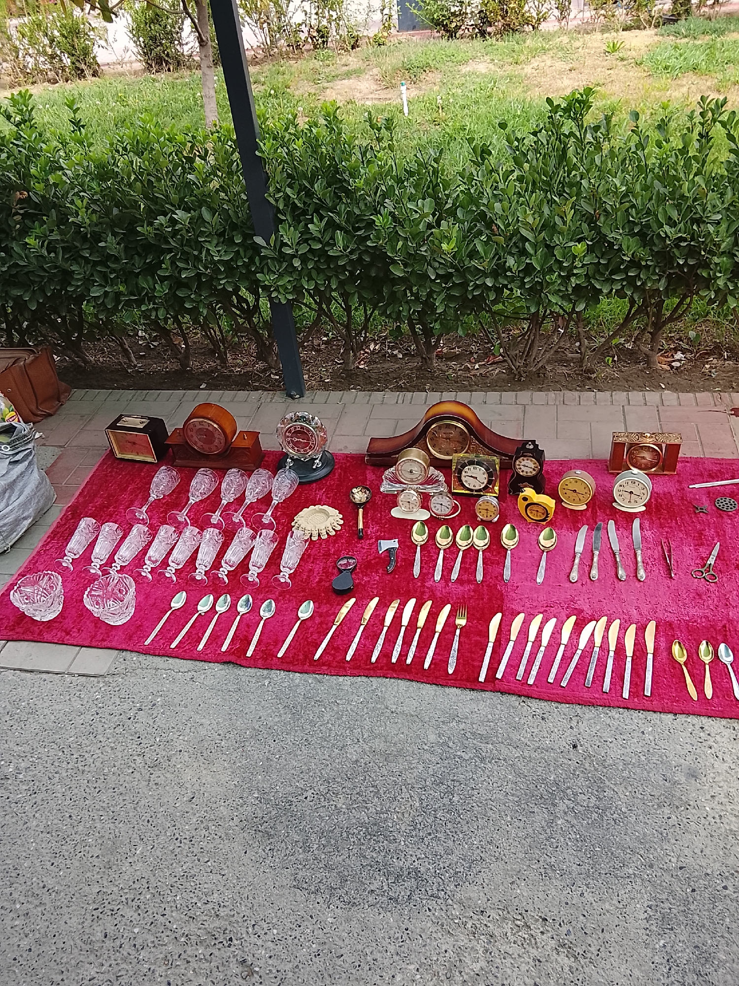 An outdoor market stall in Fergana City, Uzbekistan, displays a variety of antique and collectible items laid out on a red cloth. The arrangement includes several ornate clocks, including mantel clocks and alarm clocks, some with visible clock faces. There are also numerous crystal glasses, some in rows and others in small bowls. A collection of spoons and knives, some appearing to be silverware, are neatly arranged. A decorative piece resembling a small urn or bowl with intricate carvings is also present. Smaller items like tweezers and scissors are visible. The background consists of green bushes and a paved ground. The lighting suggests daytime.
