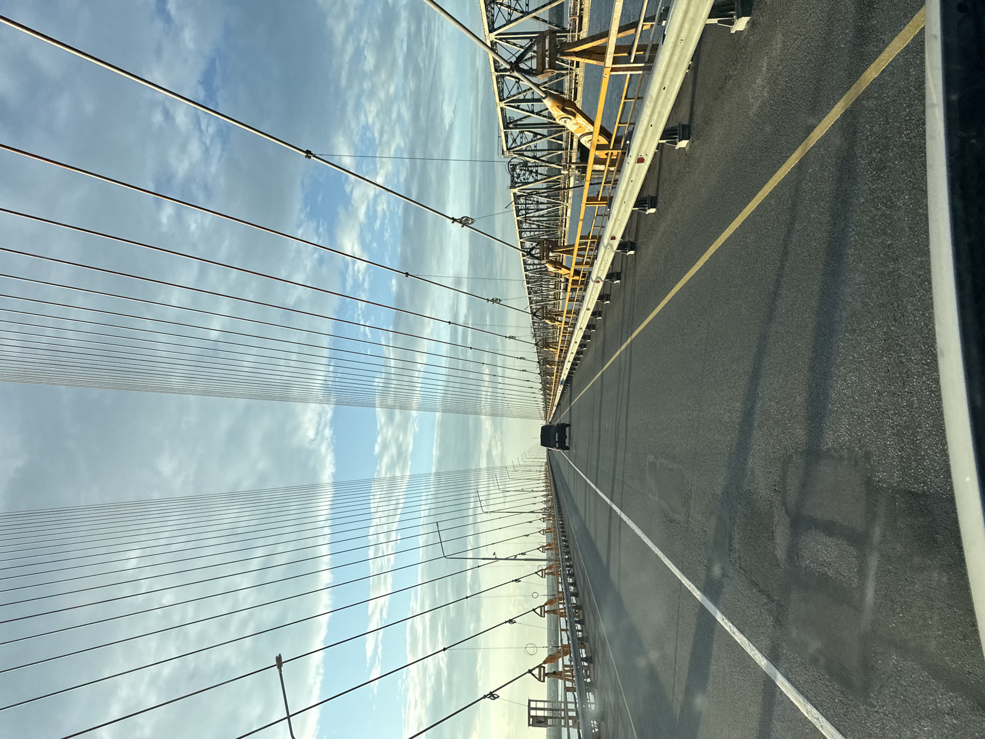 The image shows a perspective from inside a vehicle traveling on a cable-stayed bridge. The road is paved and has a white line marking the lane. A dark-colored SUV is driving ahead in the same lane. The bridge has tall pylons from which numerous cables extend, supporting the roadway. On either side of the road, there are guardrails and lampposts. The sky is partly cloudy with patches of blue visible. The scene appears to be in an urban or semi-urban environment, given the presence of the large bridge and infrastructure. The bridge spans over water, and a metal truss structure is visible in the background, suggesting a complex transportation network. The overall impression is one of modern infrastructure and transit.
