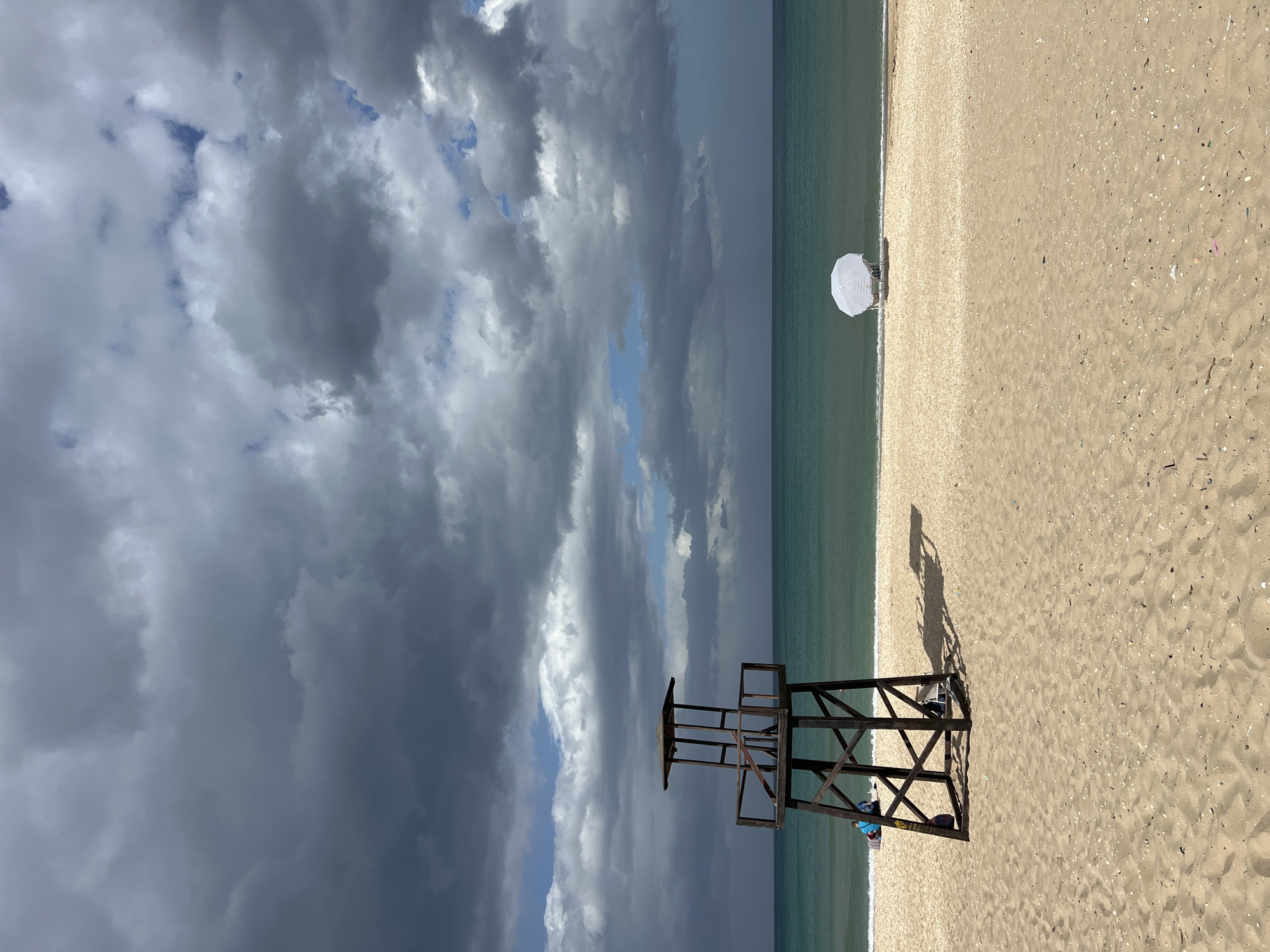 This is a photograph of a beach scene in M'Diq, Morocco. A wooden lifeguard tower stands on the sand, casting a shadow. An umbrella is visible on the beach near the ocean. The sky is filled with dark, ominous clouds. The ocean is a greenish-blue color. The sand is light brown.