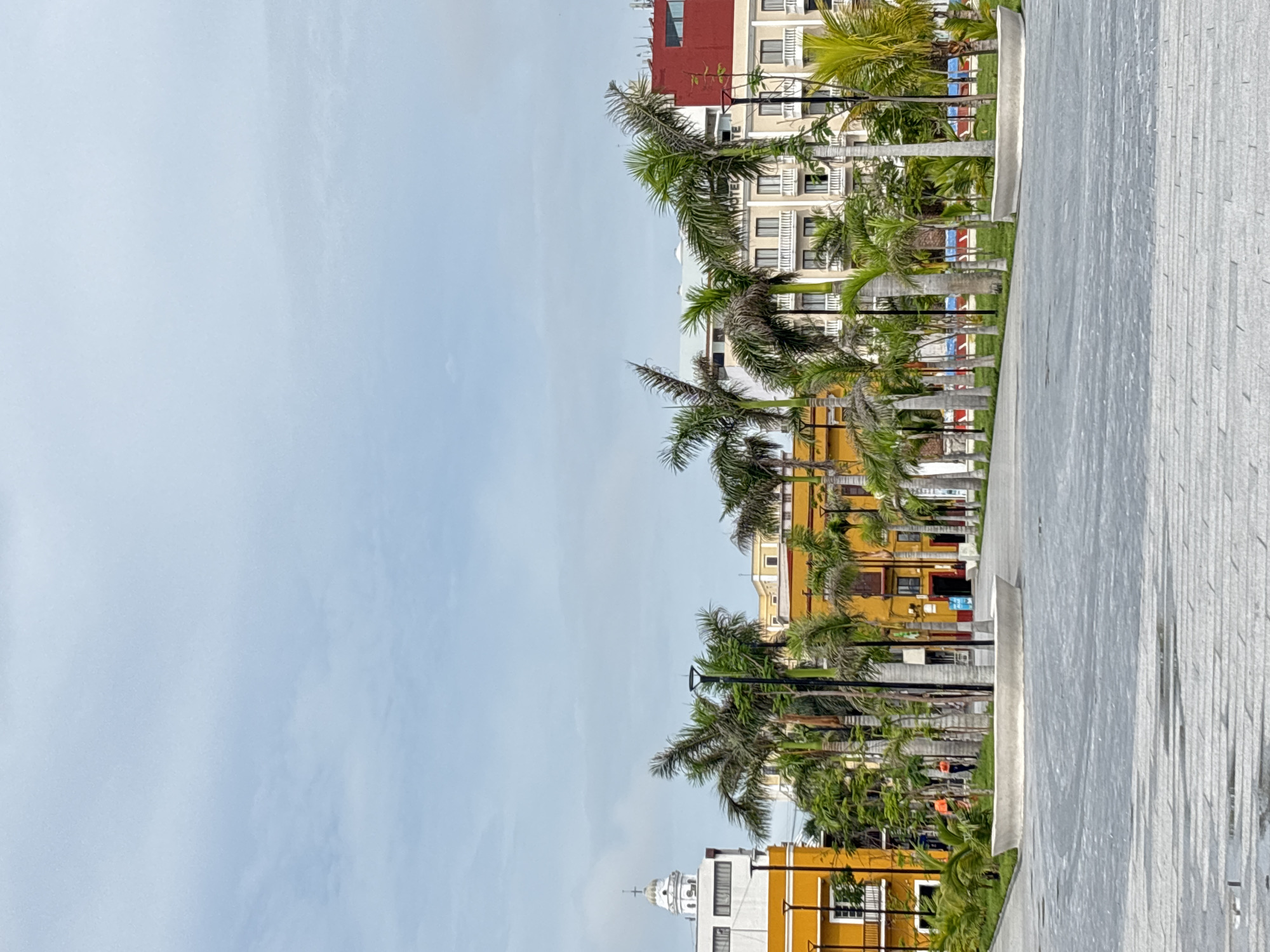 The image depicts a public plaza in Veracruz City, Mexico, likely near the Malecón waterfront. The foreground shows a paved area with a low, curved concrete planter. A row of tall palm trees lines the plaza, separating the paved area from the buildings beyond. The buildings are a mix of architectural styles and colors, with yellow facades and white trim being prominent. One building on the left has a white dome with a cross, suggesting it might be a church or other significant structure. To the right, a larger building with multiple stories and balconies appears to be a hotel. The sky is overcast with a pale blue hue. Two individuals wearing bright orange tops are visible in the middle ground, walking. The overall impression is one of a well-maintained urban public space with tropical vegetation and a blend of historic and modern architecture.