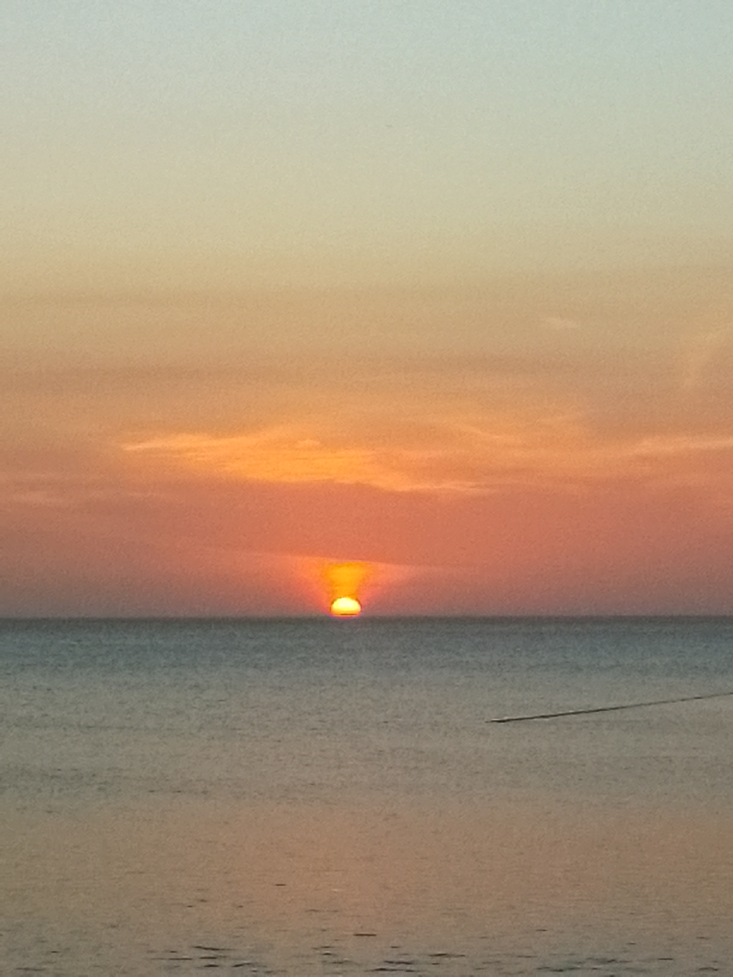 This image captures a tranquil sunset over the sea in Juan Griego, Venezuela. The scene is dominated by the horizon line, dividing the sky and the water almost equally.
The setting sun, a luminous yellow-orange disc, is partially submerged below the horizon, appearing flattened and slightly elongated. Its brilliant light creates a warm glow around it. The sky above presents a gradual transition of colors, starting with a pale, soft blue-grey at the top, fading into a hazy yellow, and then deepening into rich shades of orange and reddish-orange closer to the sun and horizon. Subtle, wispy cloud formations are visible as faint textures within the orange segment of the sky.
The sea, occupying the lower half of the image, is a calm, deep blue-grey, gently reflecting the soft, warm tones of the twilight sky. Its surface shows very slight ripples, suggesting still waters. A faint, thin, dark line or object stretches across the water in the lower right quadrant, possibly indicating a distant boat's trail or another subtle surface feature.
The time of day is clearly sunset, with the sun low on the horizon, casting a peaceful and serene ambiance over the coastal landscape. The weather appears clear, with only minimal atmospheric haze contributing to the color display. There is no visible text in the image.