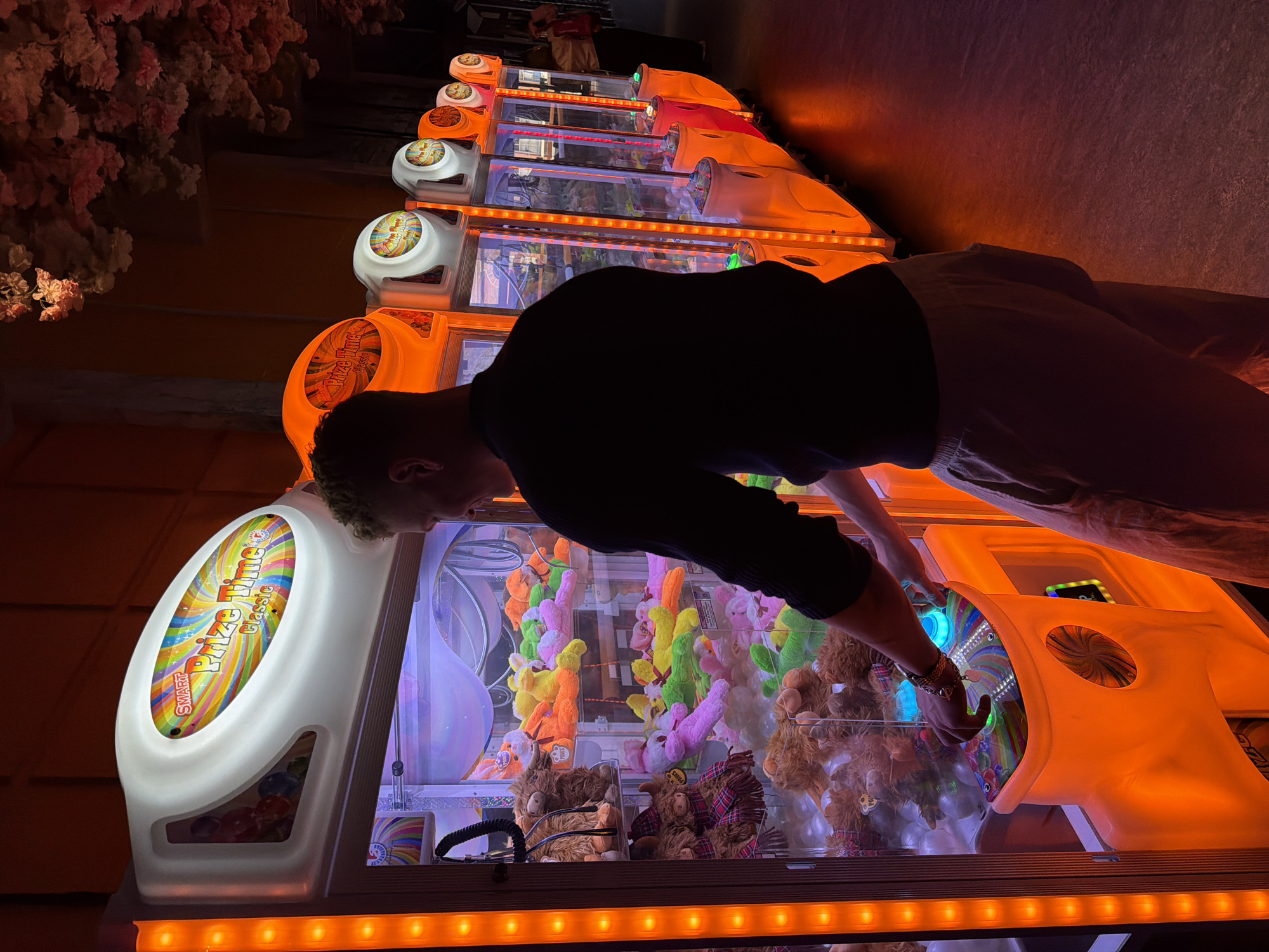 A person is standing at an arcade prize game cabinet at an unidentified location in London, UK. The cabinet is labeled "Prize Time Classic," filled with a variety of stuffed animals, and illuminated with bright neon lighting. The person, wearing a dark top and light pants, is reaching into the cabinet. Additional similar game cabinets extend in a row into the background. Flowers or foliage are visible in the upper left corner.