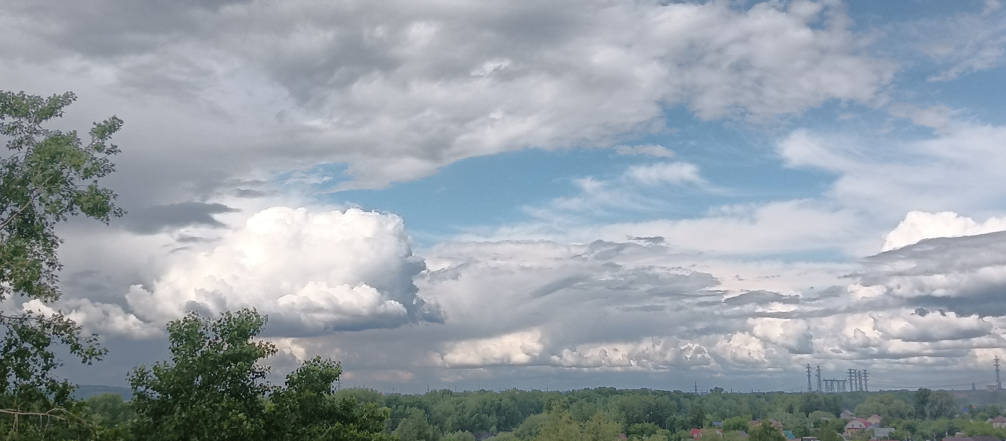 The image shows a landscape with a sky filled with clouds and patches of blue sky. Trees are visible in the foreground, with a forest spanning across the middle ground. In the background, there are electrical towers and some houses. The location appears to be a suburban or rural area. The city is Unknown, Unknown.
