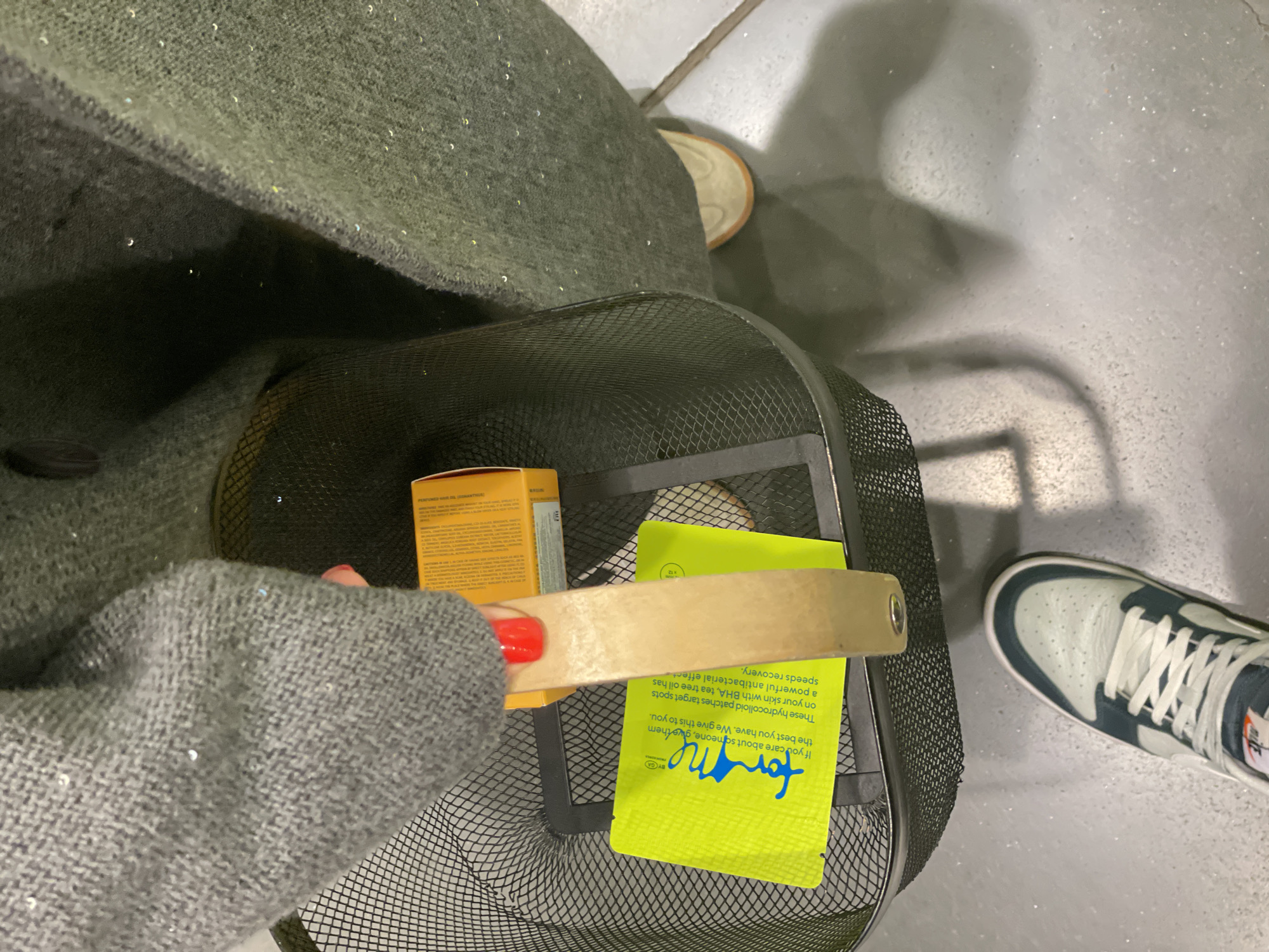Here is a detailed description of the image:
A high-angle shot shows a person from the chest down, wearing a gray sweater and a dark green coat, standing on a gray floor. A black wire basket is in front of them, containing a box of "PERFUMED HAIR OIL (OSMANTUS THORNS)" and a bright yellow package labeled "for me BY GA." A wooden handle lies across the basket's top, held by the person's hand with red nail polish. To the right, a white and blue sneaker is visible on the floor. There is also another shoe, a tan color, behind the basket and coat. The environment appears to be indoors, likely a store or home, given the flooring and the items in the basket.