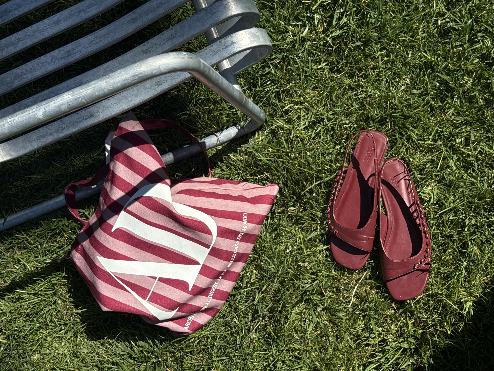 A flat lay image shows a canvas tote bag with red and pink stripes and a large white "AN" logo, lying on green grass. Next to the bag is a metal bench. Two burgundy flat shoes with decorative studs and straps are placed on the grass in the foreground. The scene appears to be outdoors in a park or garden setting. The text on the bag reads "ARCHITECTURAL DIGEST, LE PIÙ BELLE CASE DEL MONDO."