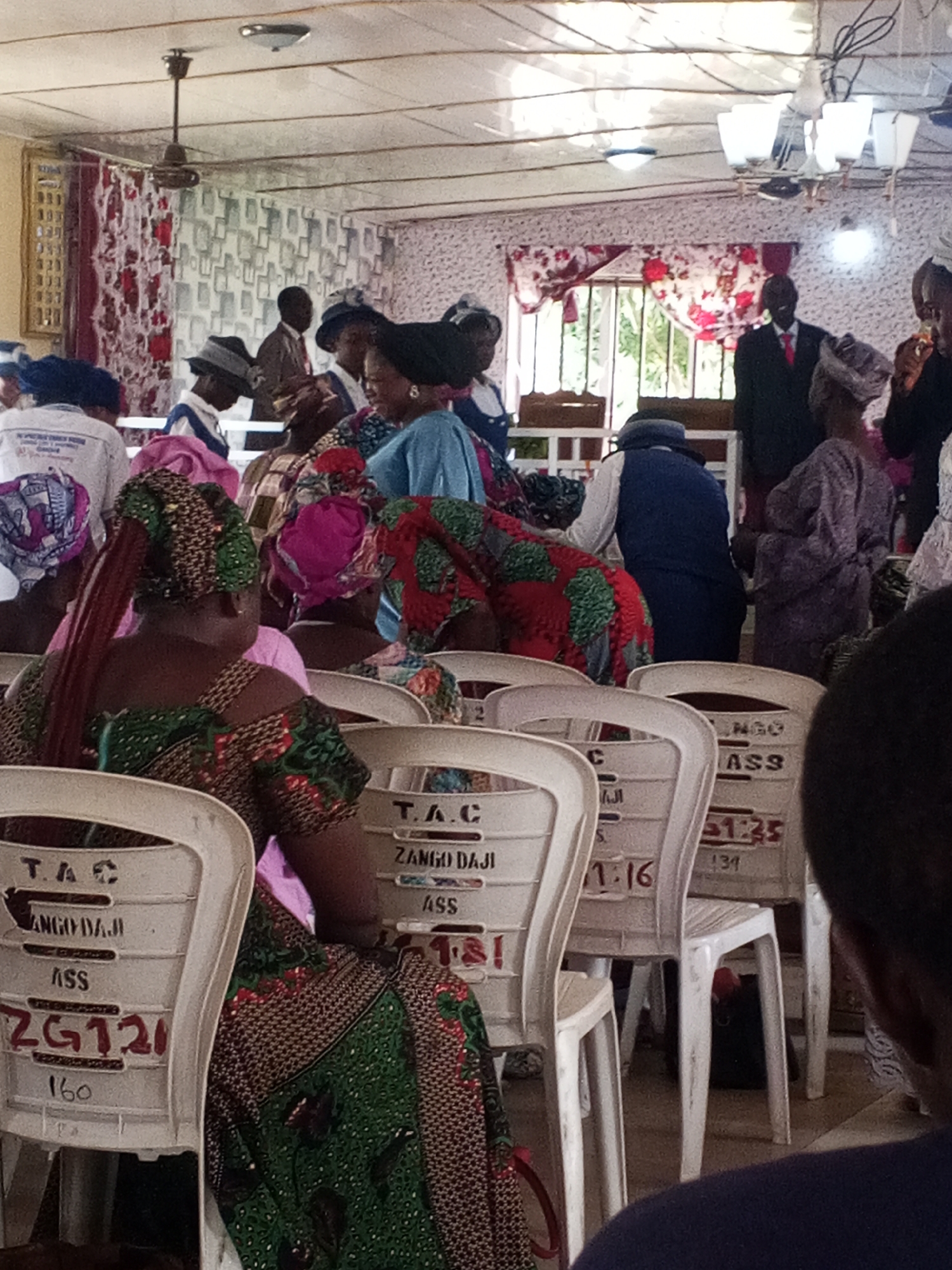 The image depicts an indoor gathering, likely a religious service, taking place in Zango-ndaji, Nigeria. The room has a simple interior with white walls and a tiled ceiling. A ceiling fan is visible, along with strip lighting. The environment is furnished with rows of white plastic chairs, some of which have labels like "T.A.C", "ZANGO DAJI", "ASS", and numbers such as "ZG121", "160", "31:25", and "134" printed on them.
Several individuals are present, all appearing to be adults. Many are wearing headwraps or hats in various colors and patterns. The individuals in the foreground are women, facing away from the camera and seated in the chairs. They are dressed in traditional Nigerian attire, characterized by vibrant fabrics and elaborate head coverings. One woman in the immediate foreground wears a green and red patterned outfit with a matching headwrap, and has long braided hair. Another woman to her left wears a blue garment and a black headwrap.
In the background, a man in a suit and red tie stands near a window, appearing to be leading or officiating the gathering. Other individuals are visible further back, some also in traditional attire, and some in more formal wear. The lighting appears to be a mix of natural light from windows and artificial lighting from the ceiling fixtures. The overall atmosphere suggests a communal event, possibly a church service or a similar religious assembly, given the setting and attire.