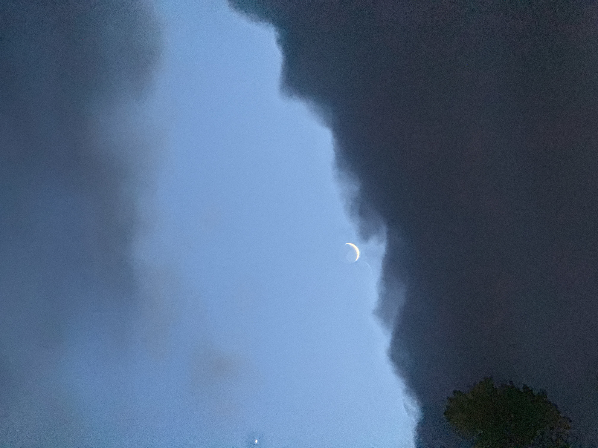 The image shows a crescent moon in a blue sky, partially obscured by dark, ominous clouds. A patch of tree leaves is visible at the bottom right corner. The location and city are not discernible from the image.