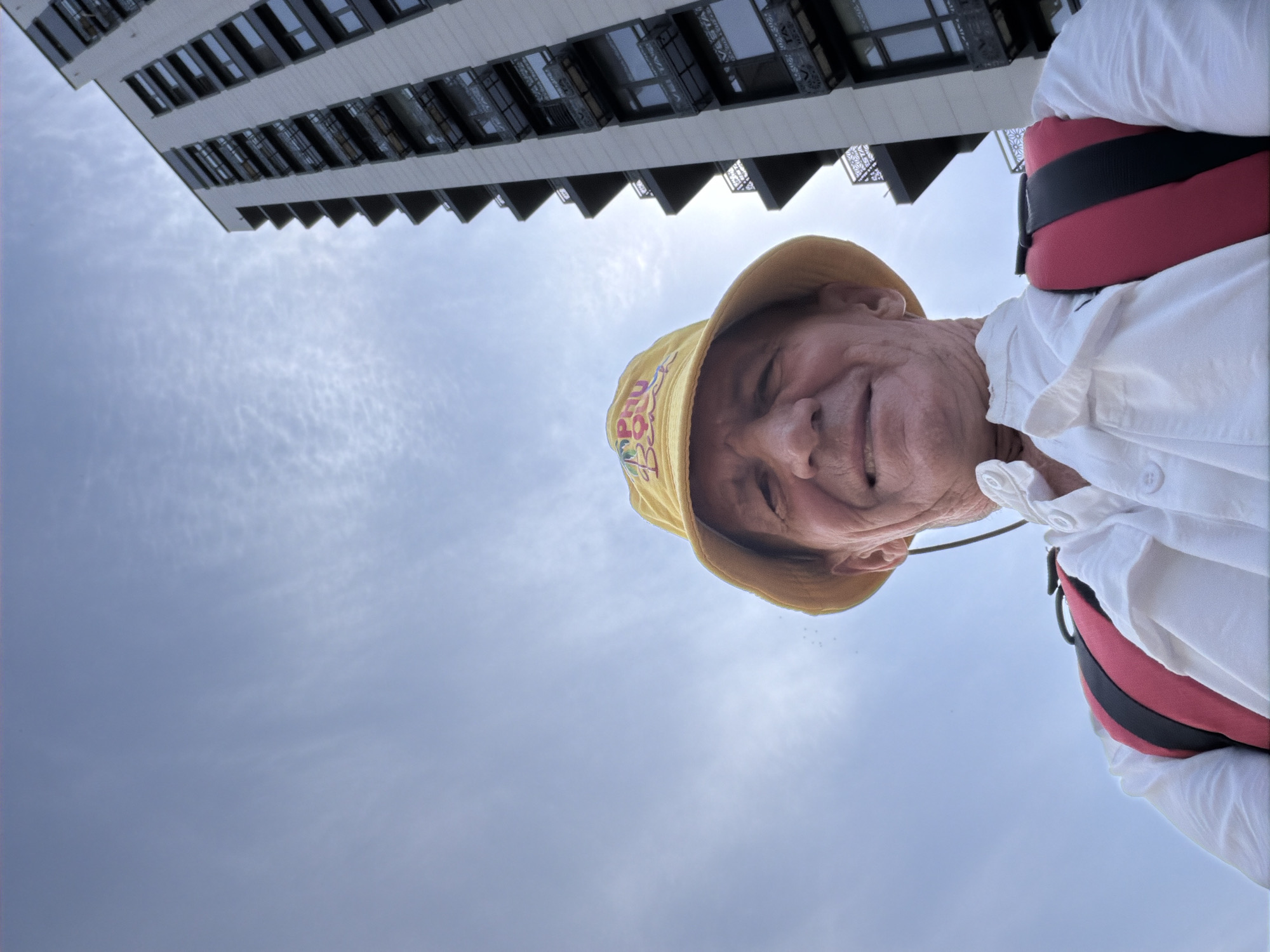 A low-angle, medium shot captures an elderly Caucasian man, facing the camera and smiling. He wears a yellow bucket hat with colorful embroidery that reads "Phu Beach". He is dressed in a white button-up shirt and a red and black backpack strap is visible over his left shoulder. Behind him, a modern, high-rise apartment building dominates the right side of the frame. The building is light-colored with numerous windows and balconies. The sky above is a pale blue with wispy white clouds. The scene appears to be outdoors in an urban environment.