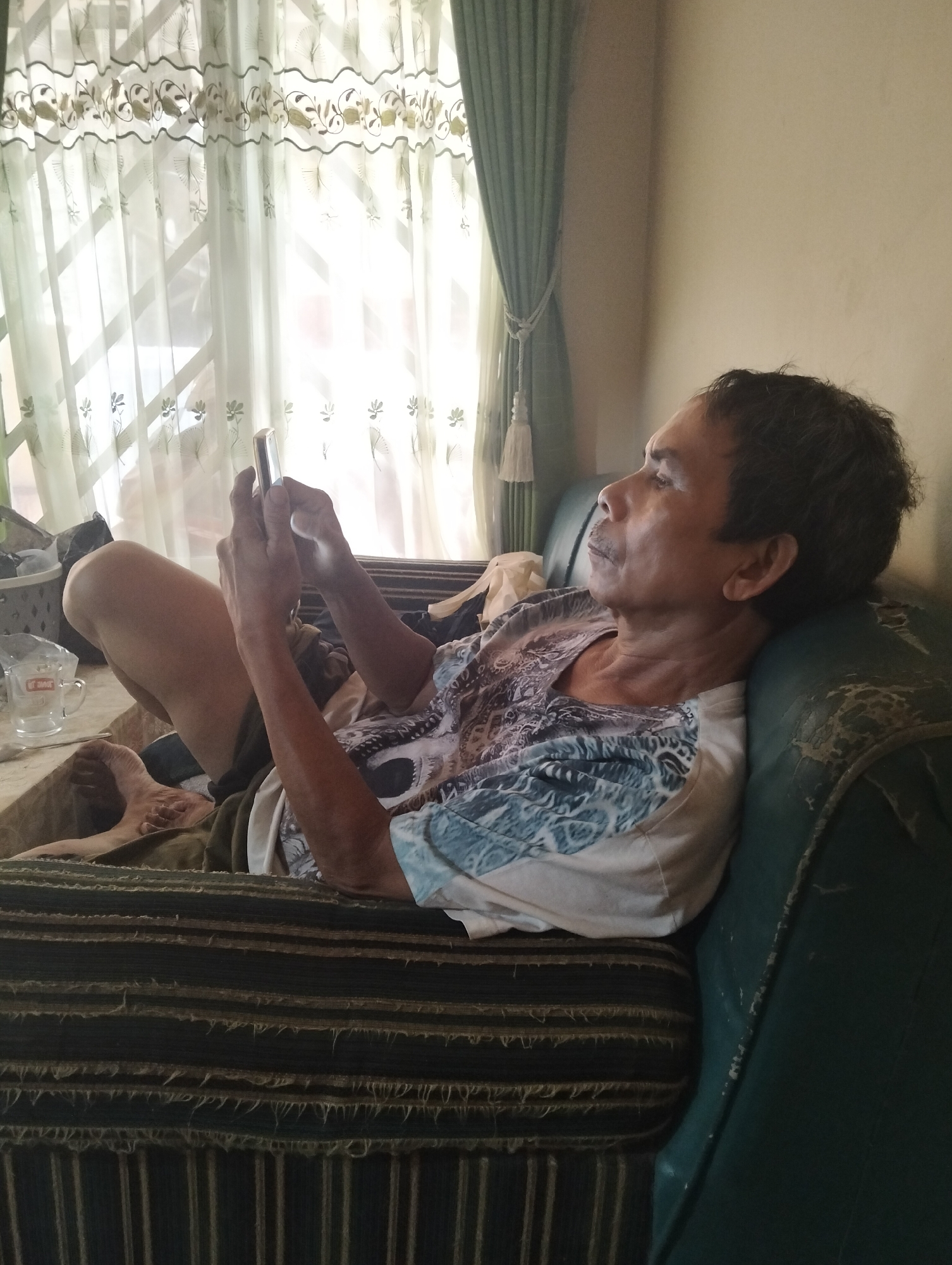 An elderly Indonesian man sits on a couch, looking at his smartphone. The couch is covered in a patterned fabric with horizontal stripes. Behind the man, a window is covered by sheer curtains with floral embroidery. The room appears to be a living room. The man is wearing a short-sleeved shirt with a blue and white pattern. His right leg is bent, and his left arm is resting on his knee. His phone is illuminated by its screen, casting a glow on his hands and face. The ambient light suggests it is daytime, and the curtains are letting in natural light. The man's facial expression is neutral, focused on the device in his hands. The couch is worn, with some fraying visible on the edges.