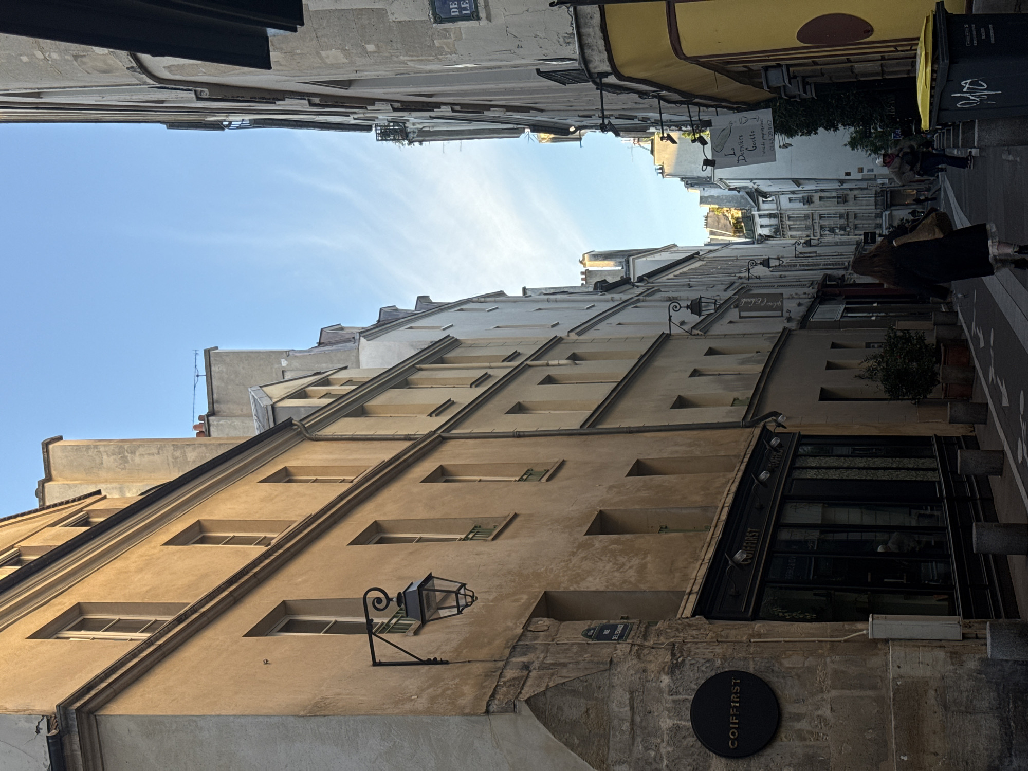 The image shows a narrow street in Paris, France, with tall buildings on either side. The buildings are primarily tan-colored with rows of rectangular windows. A black lamppost is attached to one of the buildings. A sign reading "COIFFIRST" is also visible. On the right side of the image, there is a street with people walking, along with a trash can. A sign for "La Dernière Goutte" is posted on the building. The sky is visible between the buildings. Street signs are also visible, including "Rue de l'Echaudé" and "Rue de Lévis".