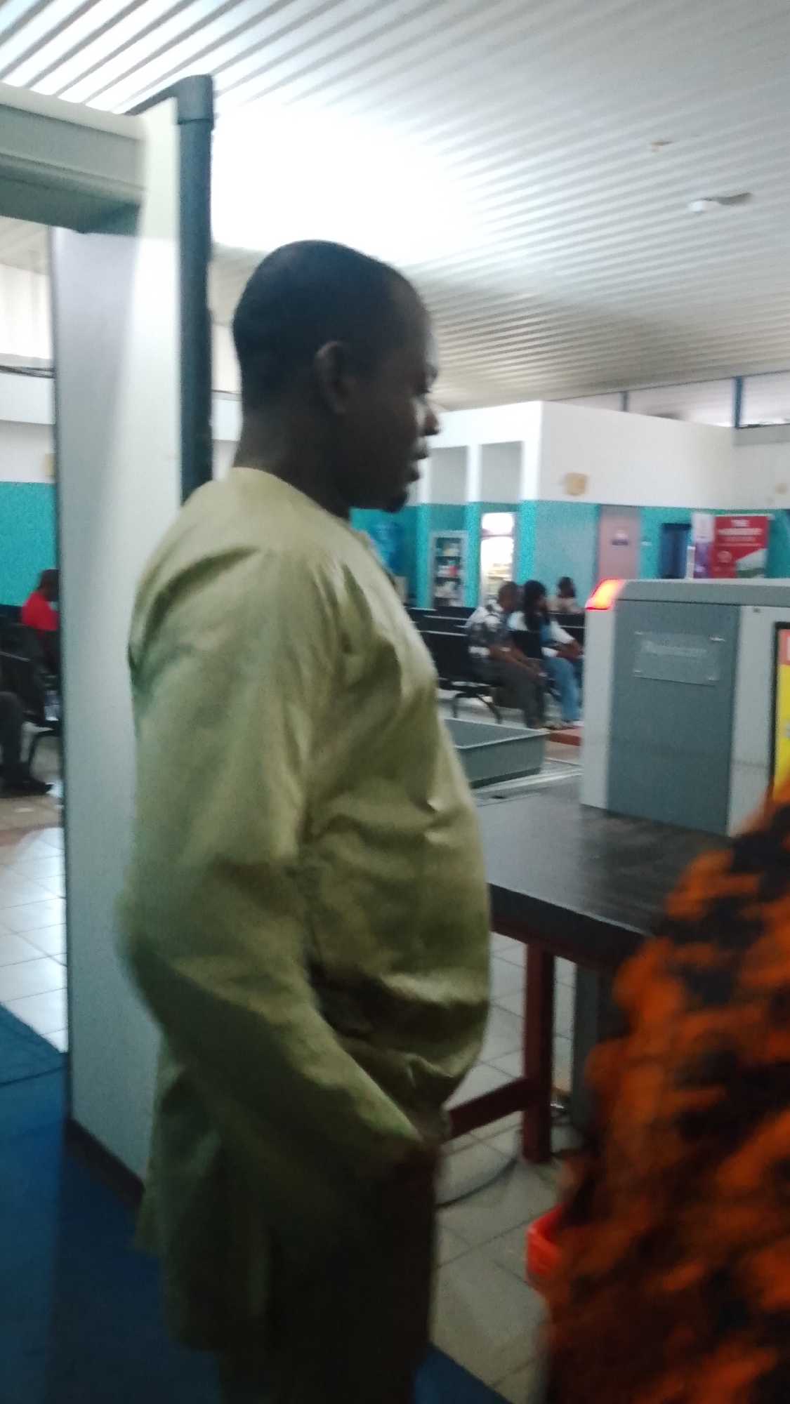 A man in a light brown traditional outfit stands to the left, facing right, with his profile visible. He is positioned in what appears to be an airport or a similar transit area, possibly within Orerokpe, Nigeria. Behind him, to the right, is a grey security screening machine with a red indicator light. In the background, several other individuals are seated on benches, waiting. The environment features tiled flooring and a suspended ceiling. The lighting suggests it is daytime, likely indoors.