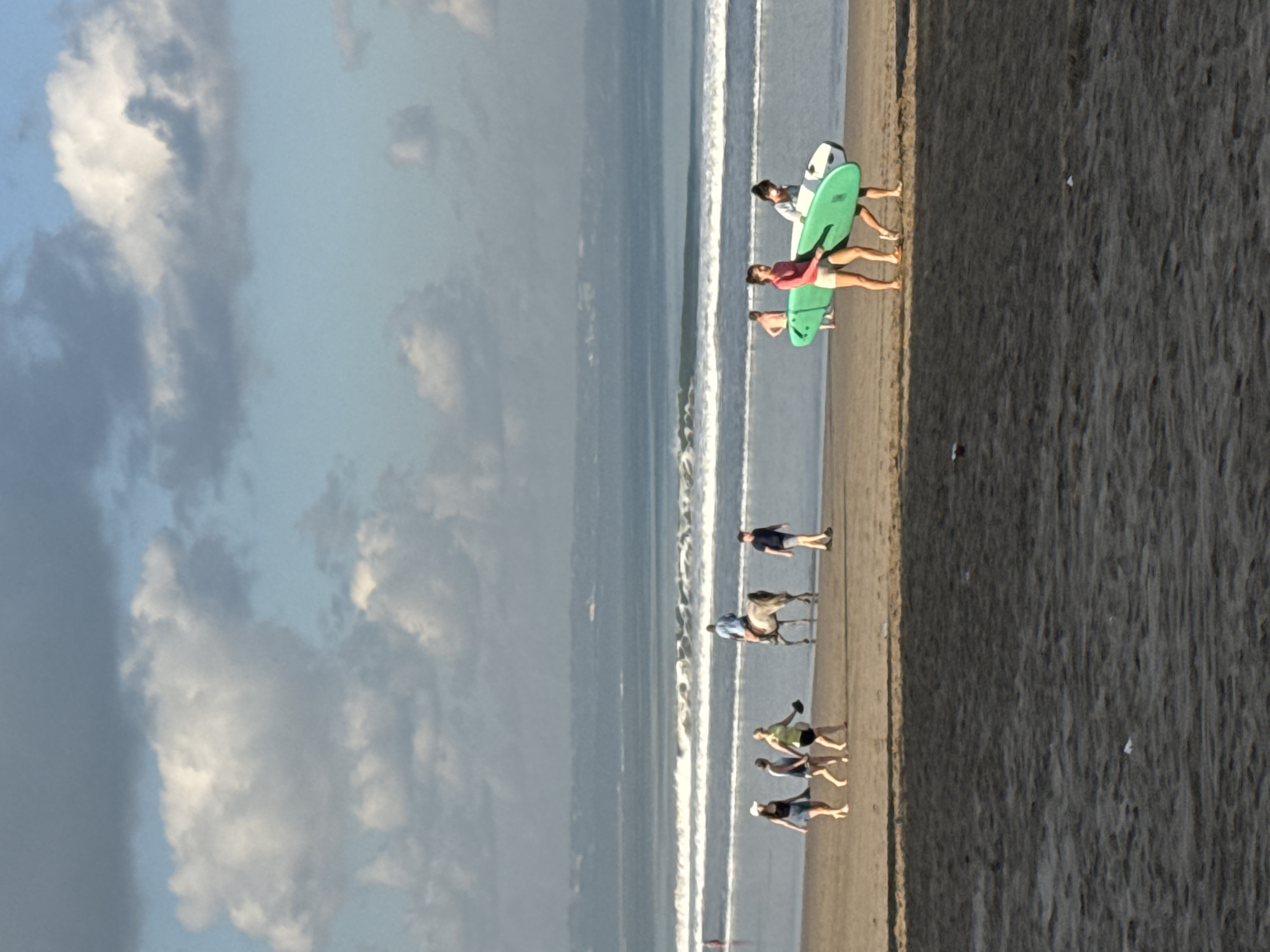This image depicts a daytime scene on a wide, sandy beach in Kuta, Indonesia, featuring the ocean, sky, and multiple individuals engaged in various activities.
The sky occupies the upper third of the frame, displaying scattered cumulus clouds of varying sizes and densities against a light blue background. Some clouds are brilliant white, while others appear denser and grayish. The horizon line is visible beneath the clouds.
The ocean occupies the middle ground, characterized by several parallel lines of small to medium-sized breaking waves with white foam crests. The water near the shore is a light blue-gray, deepening to a darker blue further out. A faint, hazy landmass or distant coastline is discernible on the horizon.
The beach itself, in the foreground and mid-ground, consists of light brown sand that appears wet near the water's edge and transitions to a darker, possibly shadowed, texture in the immediate foreground. Ripples and footprints are visible on the sand surface. Small, light-colored debris is scattered on the foreground sand.
Ten identifiable individuals are present on the beach.
* **Left group (4 people):** Four females are walking from left to right along the waterline. The leftmost wears a dark top, light shorts, and a white baseball cap. The second wears a dark top and light shorts. The third, slightly behind, wears a light top and dark shorts, holding a dark object in her right hand. The fourth is partially obscured. All are barefoot.
* **Horse and rider:** To the right of the first group, a light-colored (possibly gray or white) horse is walking left to right along the waterline, ridden by a person in a blue shirt and dark shorts.
* **Solitary male:** To the far right of the horse, a male in a dark t-shirt and light shorts is walking left to right, barefoot.
* **Surfer group (2 people):** Further right, two females are carrying surfboards, walking left to right. The leftmost wears a pink long-sleeved top and light shorts, carrying a bright green surfboard. The rightmost wears a white top and dark pants, carrying a white surfboard with green markings. Both are barefoot.
* **Partially visible male:** Behind the surfboard carriers, a shirtless male in dark shorts is partially visible, walking towards the water.
All individuals are engaged in recreational beach activities, including walking, horseback riding, and carrying surfboards. The scene is illuminated by natural daylight, casting subtle shadows on the sand.