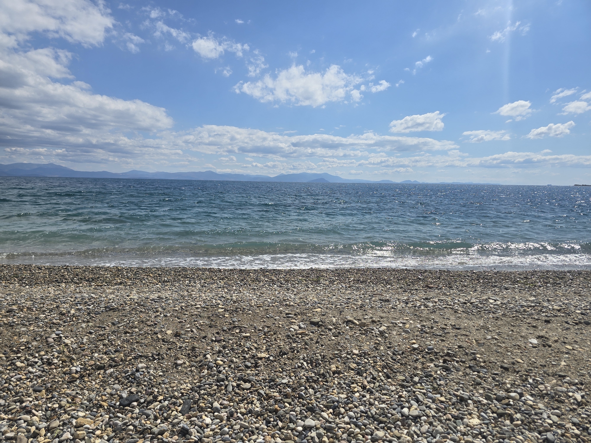 This is a wide-angle, eye-level shot of a pebbly beach in Astros, Greece. The foreground is dominated by the textured surface of the beach, composed of various sizes and colors of pebbles and sand. The middle ground features the gentle, clear blue waters of the sea, with small waves lapping at the shore. In the background, a distant mountain range is visible, silhouetted against a bright blue sky dotted with scattered white clouds. The sun is shining, creating a slight glare on the water and casting a light across the scene. There are no people or man-made objects visible in the image.