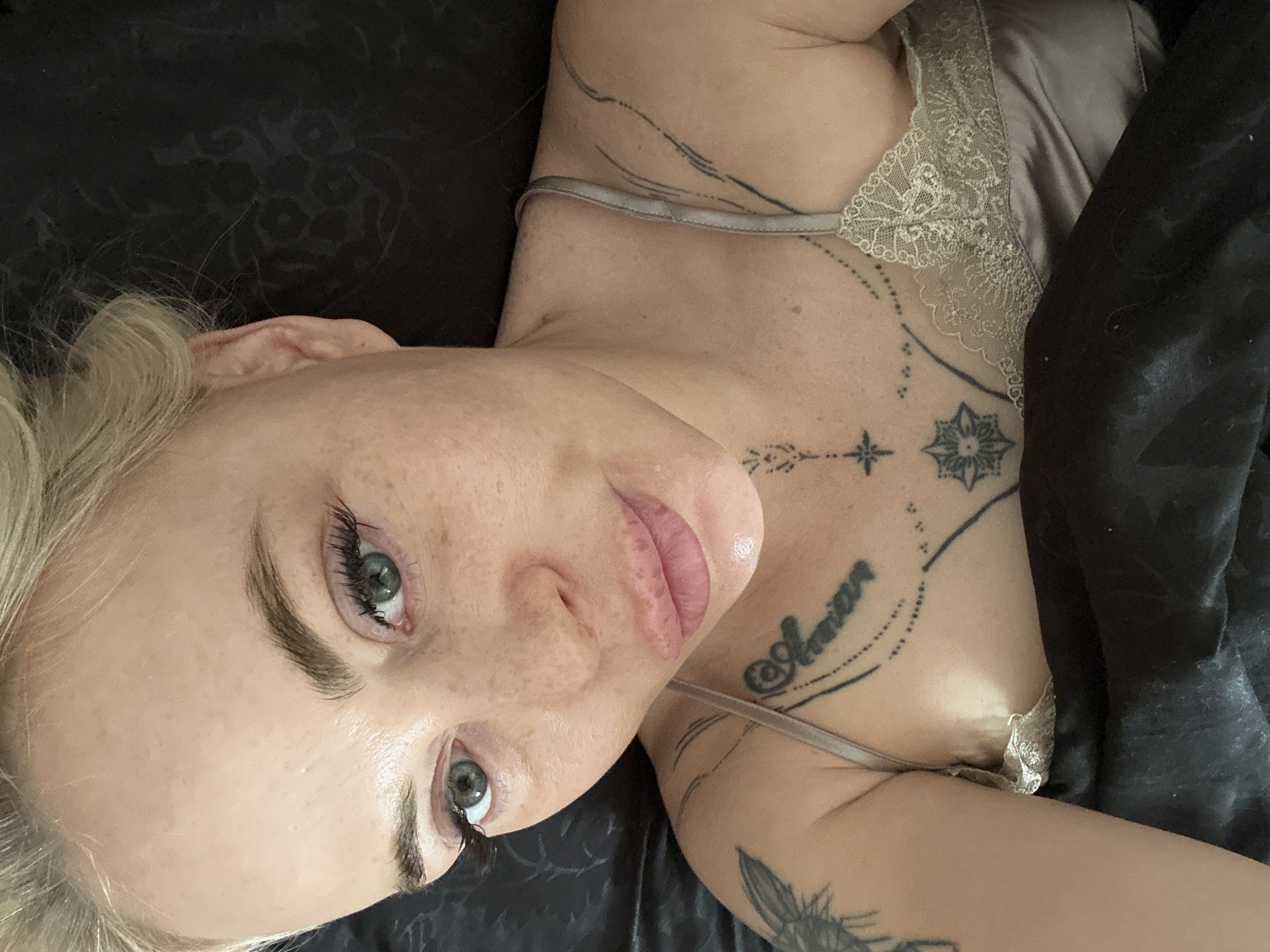 A blonde female is depicted in a high-angle, close-up shot, lying in an unknown indoor setting within the city of Unknown, Unknown. Her head is positioned on a dark, patterned fabric, likely a bedsheet or pillow. She looks directly at the camera with light green or hazel eyes, enhanced by prominent eyelashes. Her skin displays visible pores and freckles across her nose and cheeks. Her lips are full, appearing moisturized with a pink hue. Fine lines are present on her forehead. She wears a light-colored, possibly champagne or silver-grey, satin camisole featuring lace trim across the bust. Multiple tattoos are visible: a partial winged design on her upper right arm/shoulder, a script tattoo resembling "@Amona" with a dotted line on her upper chest, a four-pointed star design below her collarbone, and an intricate star or floral design on her sternum, along with abstract linear patterns extending towards her right shoulder.