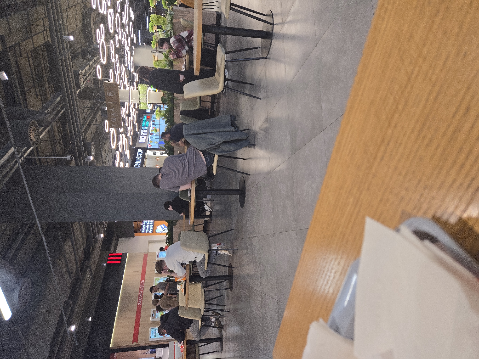 The image captures a food court area within a shopping mall, likely located in Moscow, Russia, indicated by the Russian text "ЗАКАЖИ ЗДЕСЬ" (Order Here) visible on a red banner. The scene is populated with several individuals seated at tables or standing. In the foreground, a wooden table surface with some white paper napkins is visible, suggesting a dining area.
The food court itself features a modern design with a high ceiling and industrial lighting fixtures, including circular pendant lights. Gray tiled flooring covers the main area. Several tables with chairs are arranged throughout the space.
On the left side of the image, a group of people are gathered near a counter with touch screen ordering kiosks. One person is wearing a dark hoodie, another a light brown shirt, and a third a gray t-shirt. Further to the right, a person in a white t-shirt is seated at a table, looking downwards. Another person in a dark t-shirt is visible at a table to the right of the central area, and a person in a gray t-shirt is standing.
In the middle ground, a person in a gray sweatshirt is seated at a table, with a dark jacket draped over the back of their chair. A person in a black jacket and dark pants is standing near a table, facing away from the camera. To the right, a woman in a plaid shirt is seated at a table with another person who is mostly out of frame.
The environment includes greenery in planters, contributing to a more pleasant ambiance. Various signs with text, some of which appear to be in Cyrillic script, are present on the walls and hanging from the ceiling, indicating different food establishments. The overall impression is a busy, contemporary public dining space.